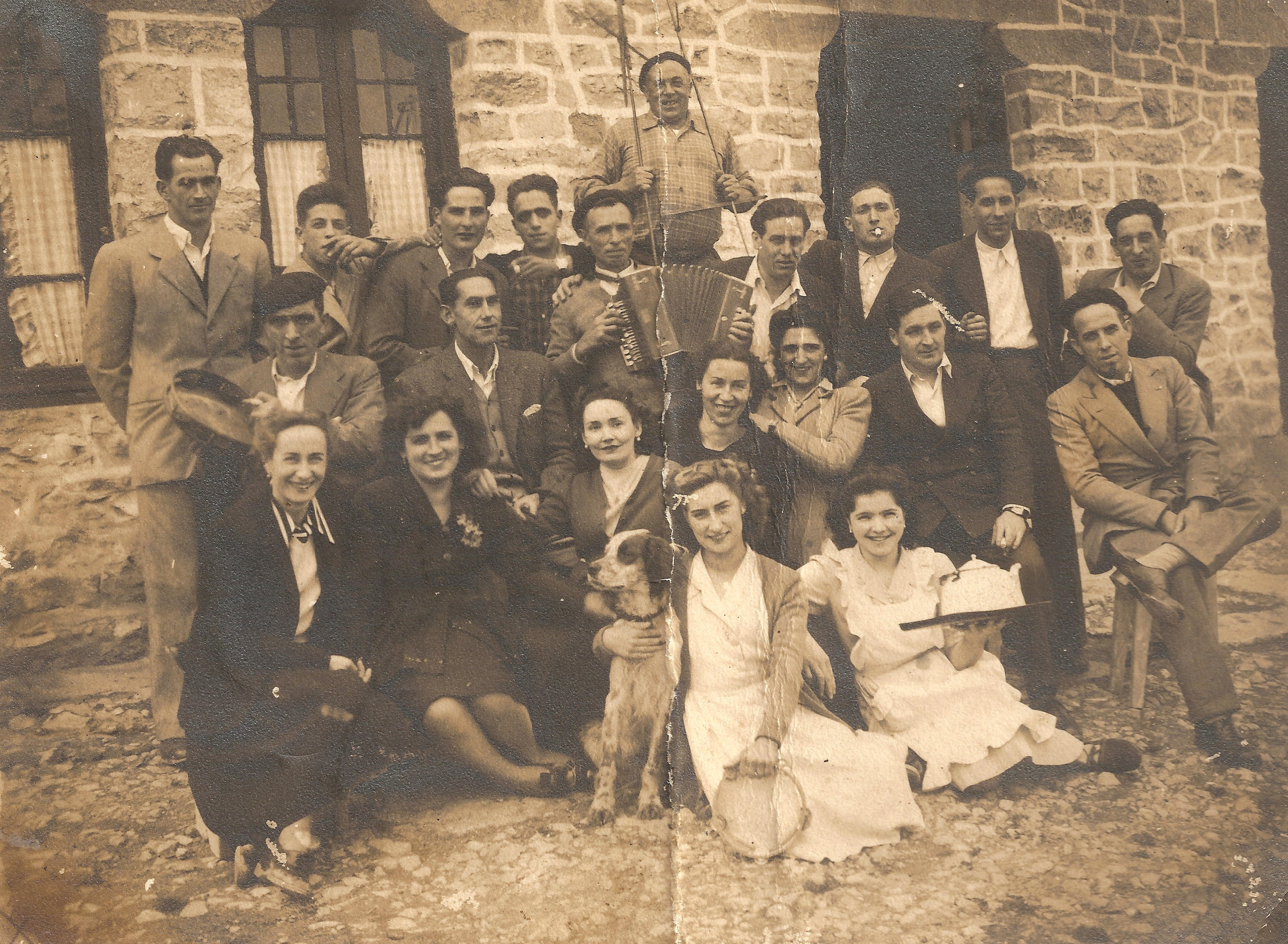 Se ve una fotografía en tonos sepia, tiene un pliegue a la mitad de haber estado doblada. Hay un grupo de trabajadores/as, unos veinte más o menos. Los hombres van vestidos en traje, y los que no llevan camisa y chaqueta. Hay un perro en la fotografía. Las mujeres visten de calle con la vestimenta típica de la época. Uno de los hombres lleva un pandero y otro una trikitrixa. Algunos hombres llevan txapela y las mujeres vestido. Parece que el ambiente es bueno, están sonrientes. 