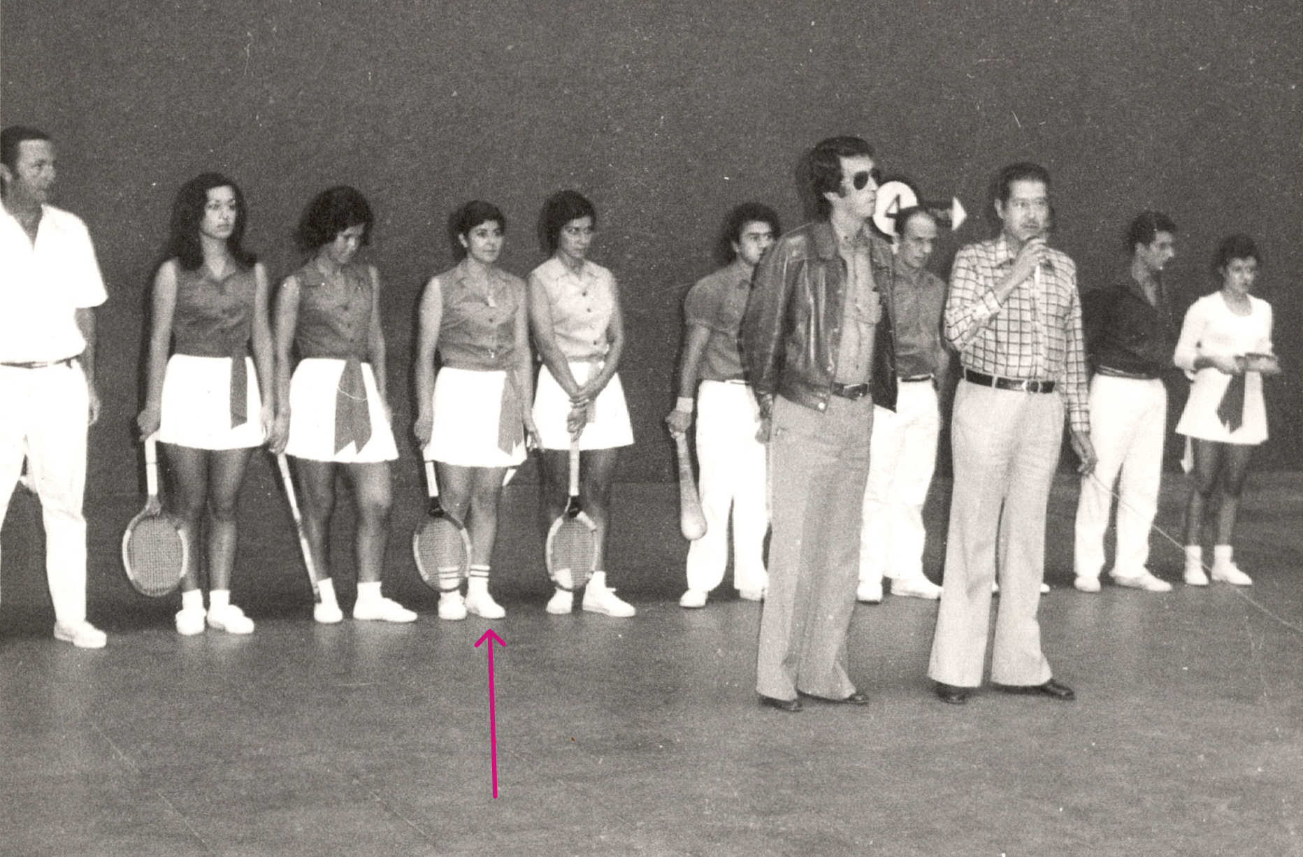 Varias raquetistas (mujeres) y palistas (hombres) están contra la pared lateral del frontón con la vestimenta tradicional del juego. Frente a ellas y ellos hay dos hombres vestidos de calle y uno de ellos habla frente a un micrófono. 