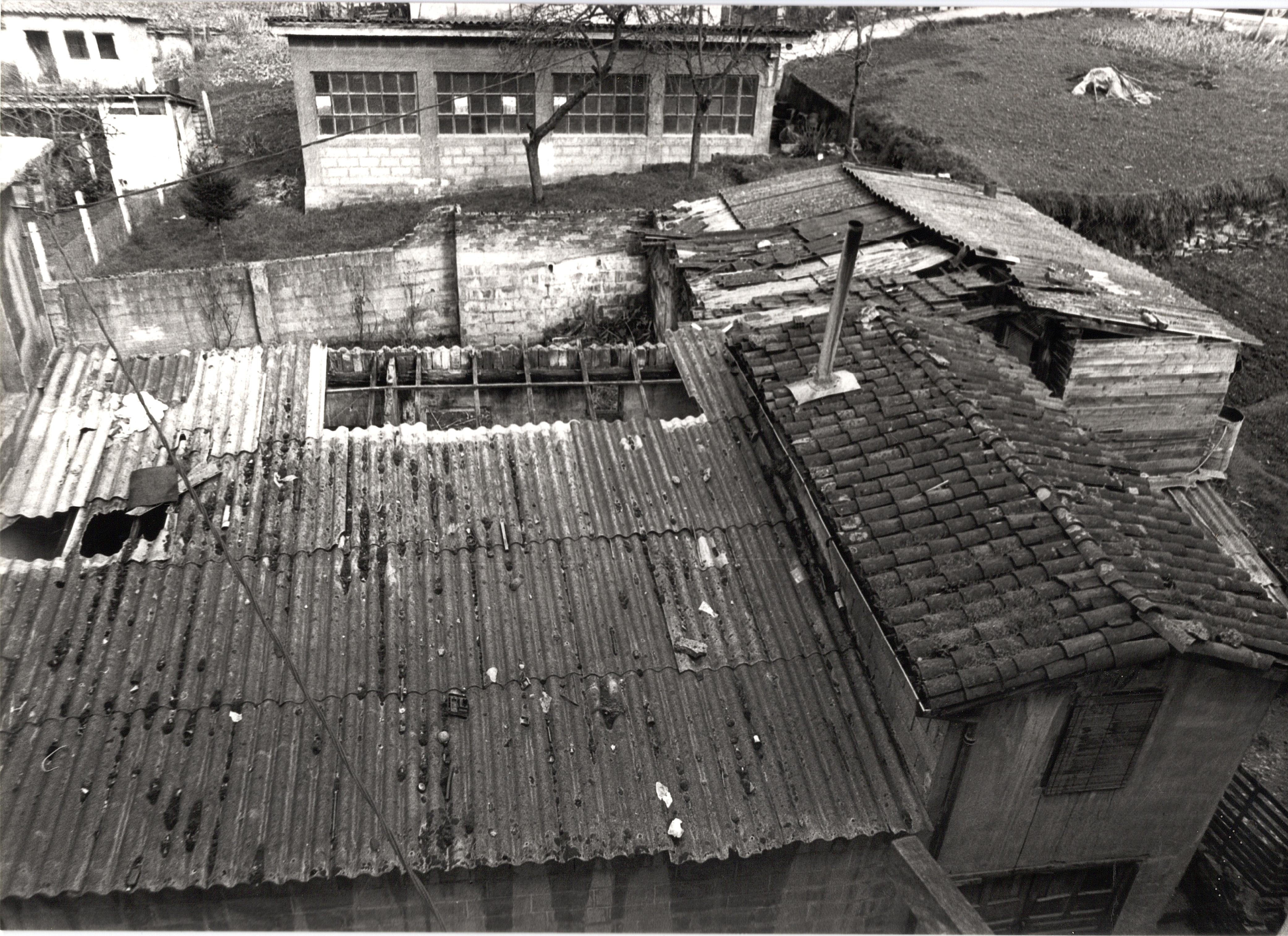 Se ven los tejados de unas chabolas, también el tejado de un edificio más robusto, con un tejado de teja y una pequeña chimenea de tubo metálico. A la derecha del conjunto de estructuras hay una parcela de hierba con una pequeña huerta. La fotografía es en blanco y negro.