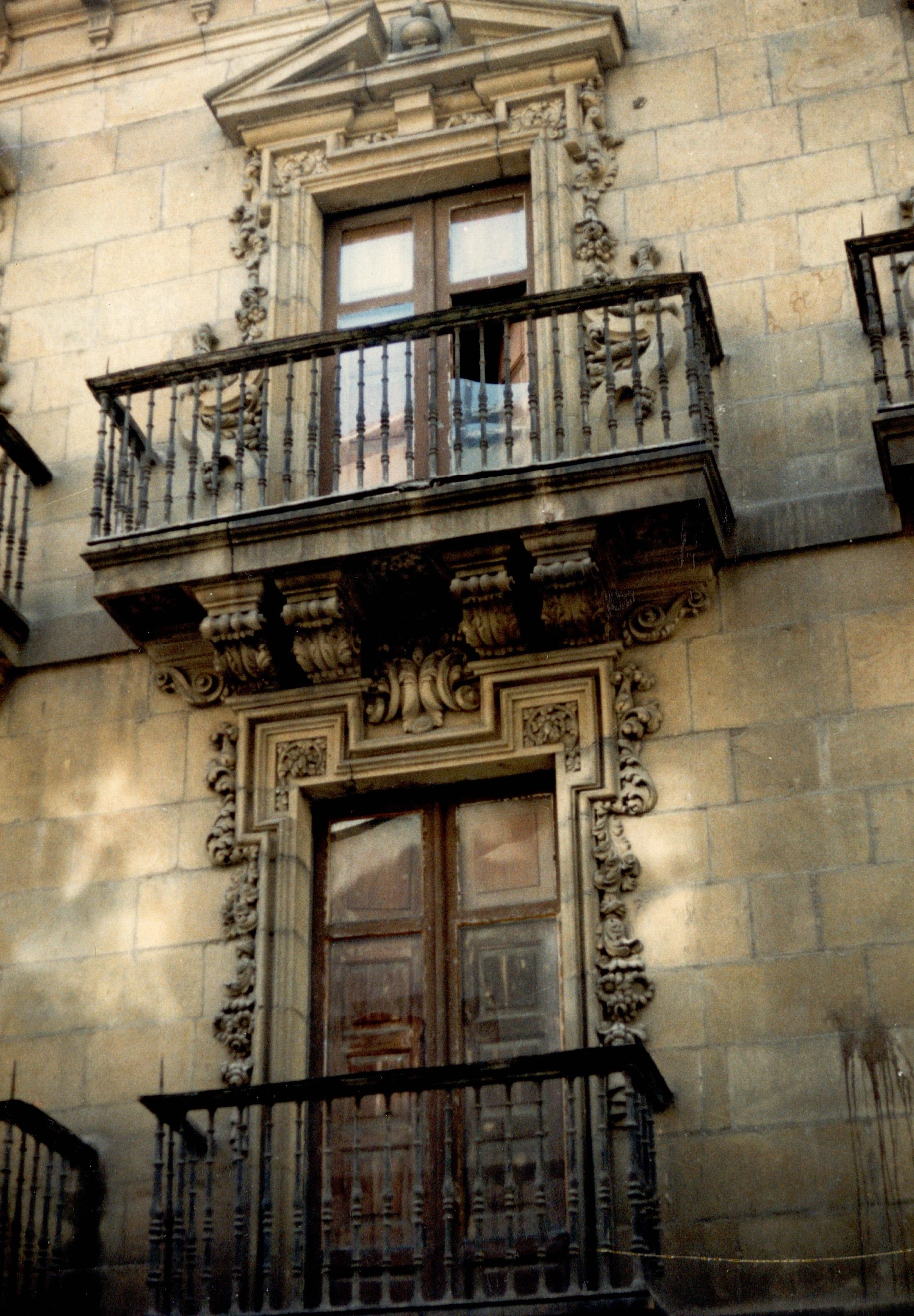 Dos balcones de un palacio, de la fachada principal. Se ven decoraciones distintas en las dos puertas, son decoraciones de piedra, de hojarisca y motivos naturales. Esos elementos decorativos rodean la puerta y la envuelven, tanto en su parte superior como en los laterales. También se ven las contraventanas de maera, algunos cristales rotos y en la fachada suciedad.