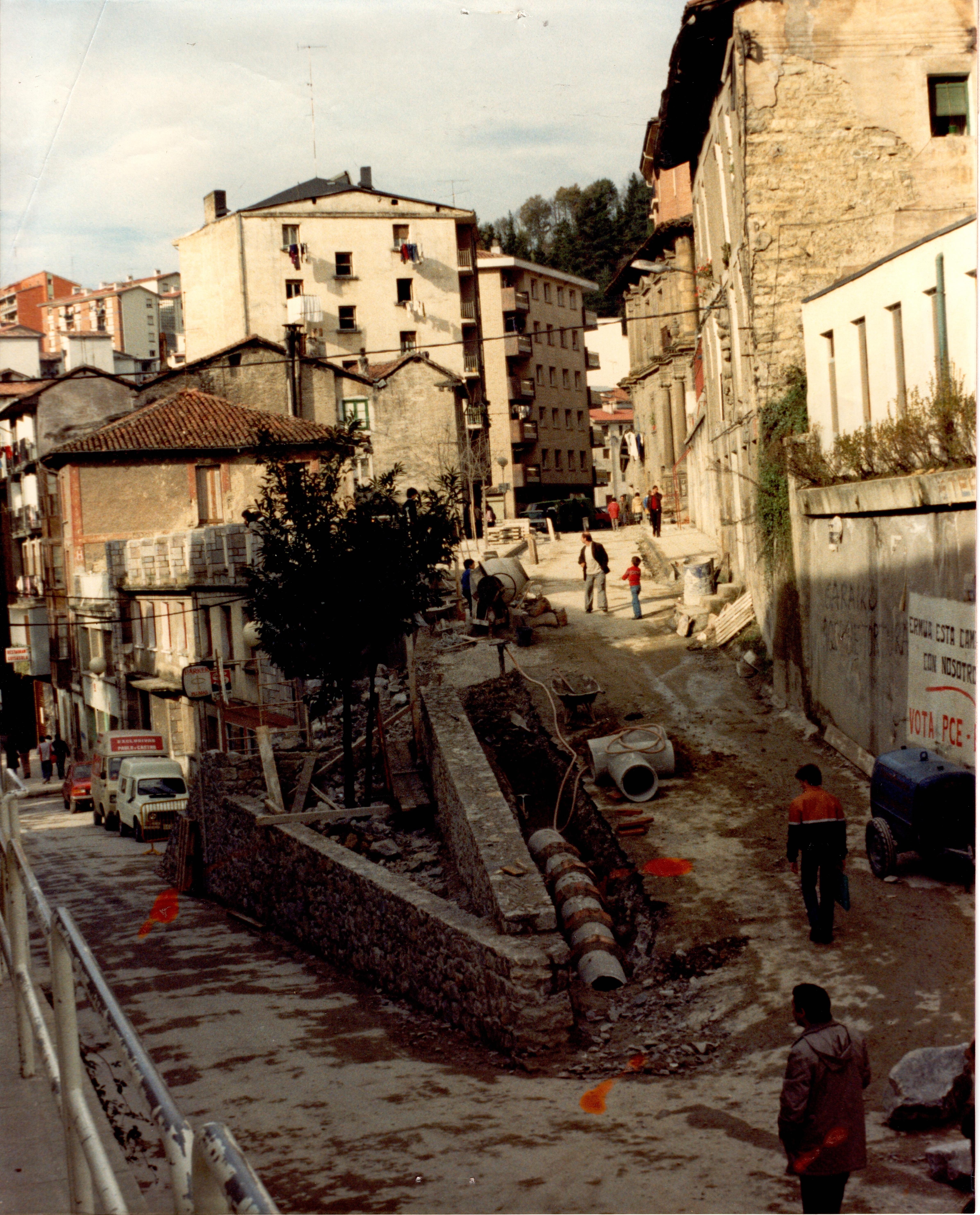 Se ven la calle Goienkale en obras, en la subida desde la calle Izelaieta. Se puede ver a dos obreros, material de obra como una hormigonera, mangueras, bidones, palets, tuberías... también se ve una carretilla y dentro de ella una herramienta de mano. El asfalto tiene tierra y agua y en esa subida a la izquierda hay una zanja donde se pueden ve las tuberías. La primera mitad de la calle o un poco más está con sombra. y el resto, más arriba, tiene sol. Más adelante se ven edificios.