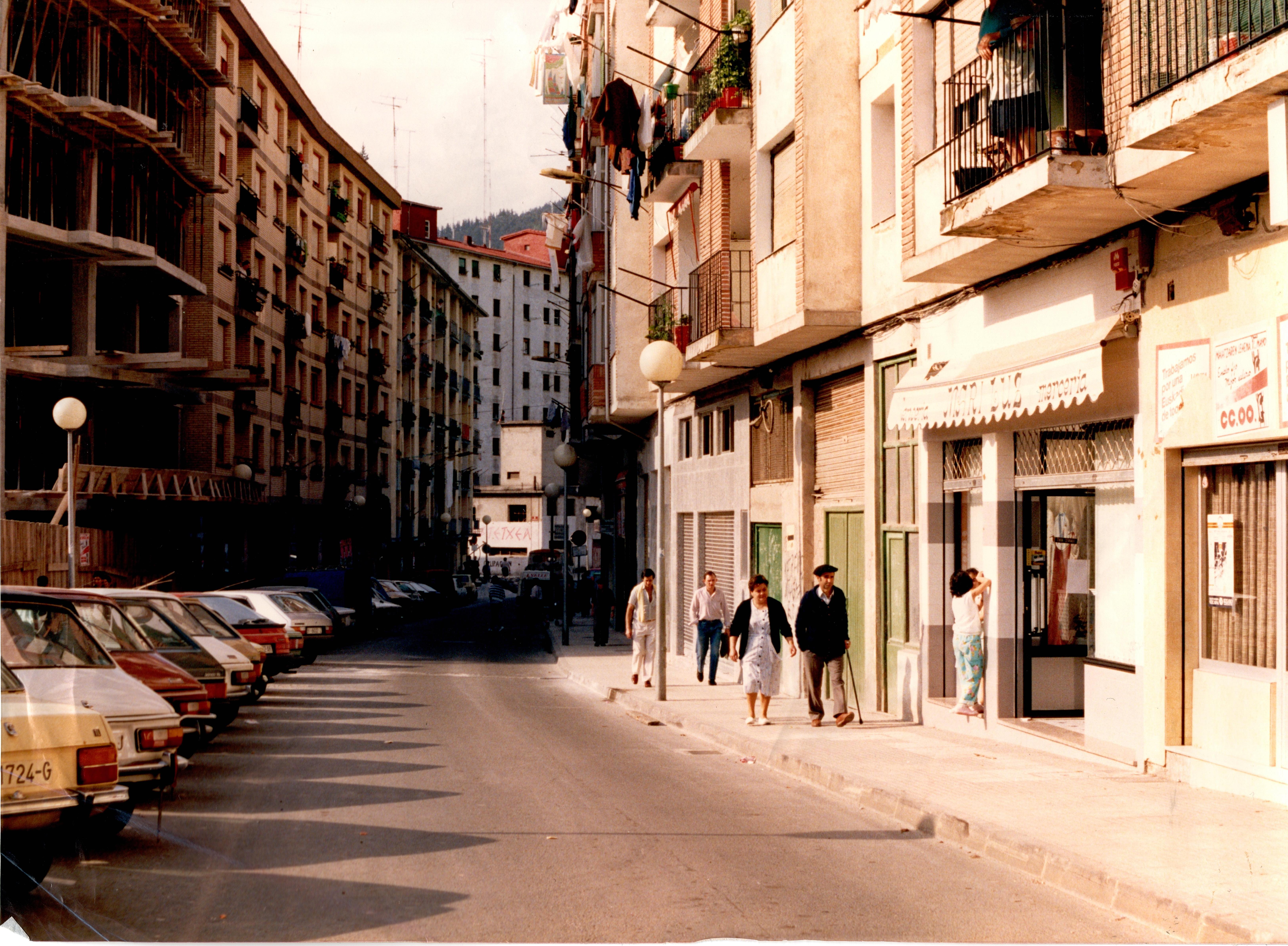 Se ve una calle con carretera en medio. A la izquierda hay coches aparcados de distintos colores en batería: color crema, blanco, rojo, negro, gris... A la derecha hay acera para peatones, en ella se ve a cuatro parejas caminando. A la derecha, bajo las viviendas está la sede del sindicado CCOO y al lado una mercería que en su toldo dice "mercería Mari Luz". Al fondo hay un edificio gris, de poca altura en el que se puede leeR "GAZTETXEA" y "OCUPACIÓN".