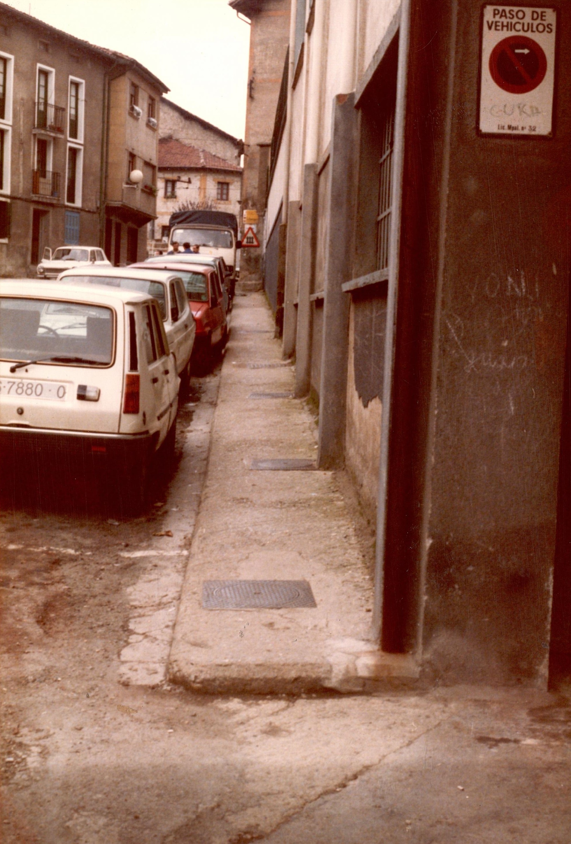 A la derecha se ve la acera junto a unos edificios. En la pared de uno de los edificios hay un cartel que pone "paso de vehículos" y una señal de prohibido aparcar con excepto. A la izquierda de la acera hay varios vehículos aparcados en paralelos, dos blancos y uno rojo y al fondo también se ve un camión. A la izquierda hay un coche en paralelo aparcado de color blanco.