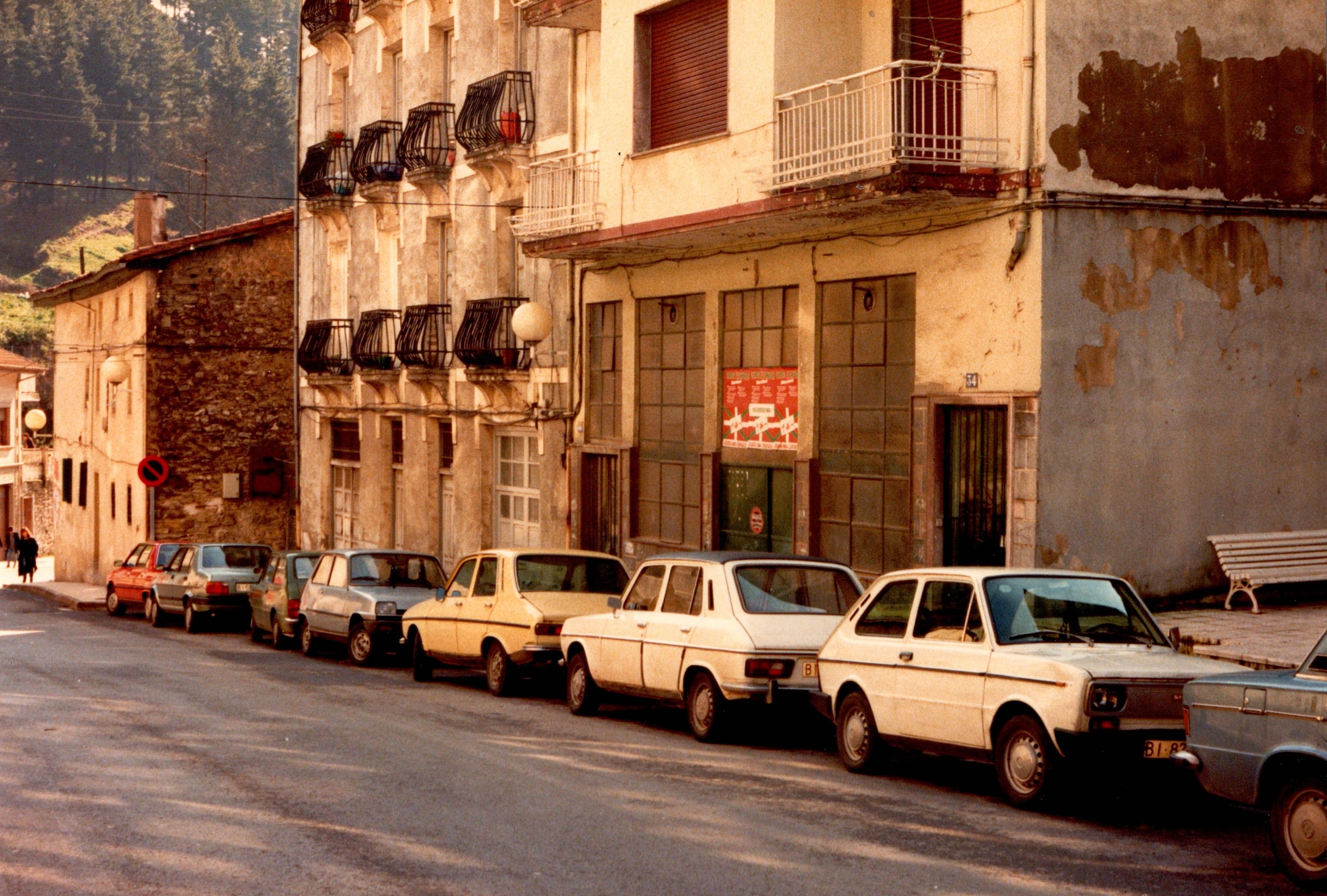Se ve la parte derecha de la calle Izelaieta, hay varios coches aparcados en paralelo de diversos colores: dos blancos, uno amarillo, dos grises, uno rojo... Hay viviendas y edificios, tres concretamente que se vean al completo.