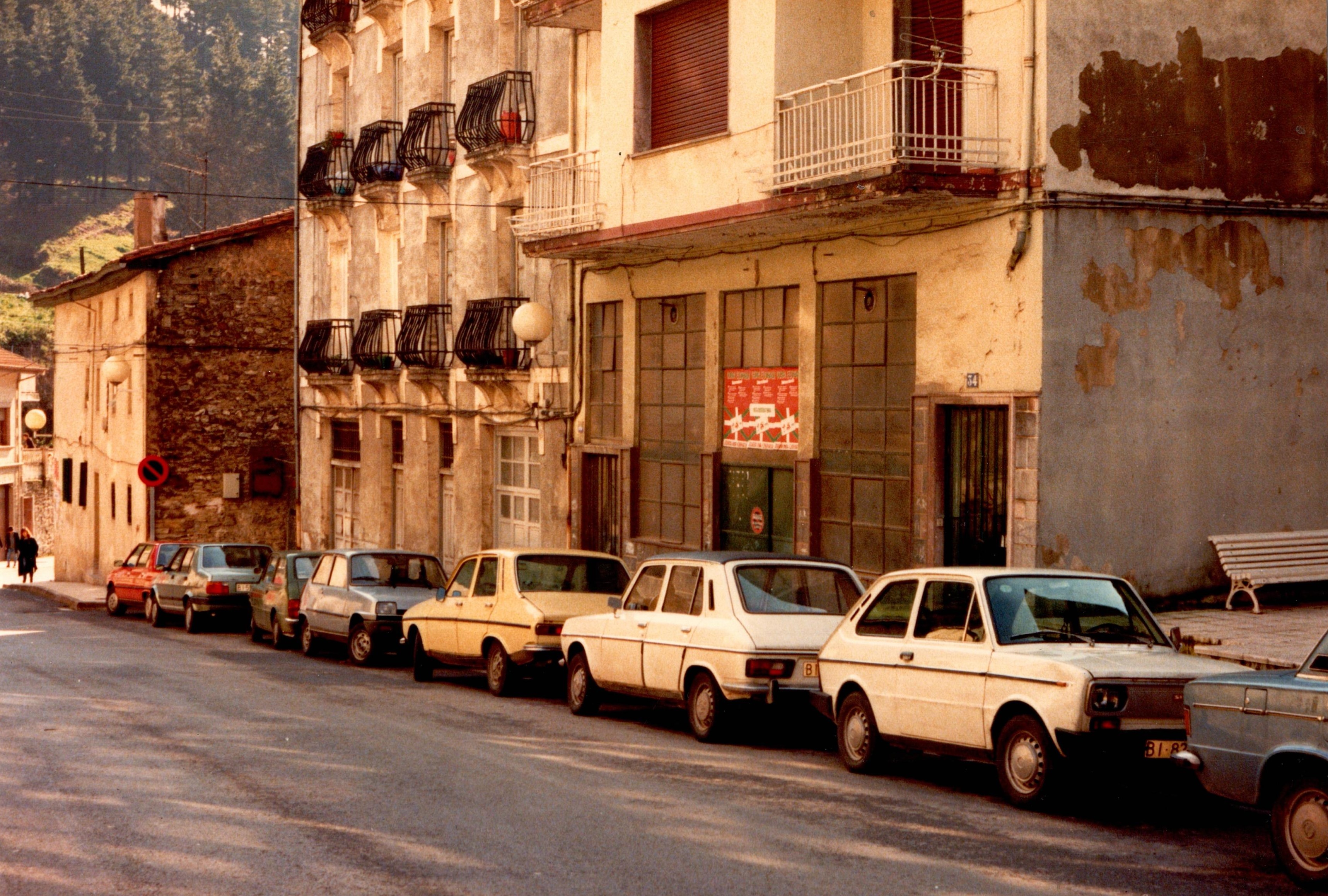 Se ve la parte derecha de la calle Izelaieta, hay varios coches aparcados en paralelo de diversos colores: dos blancos, uno amarillo, dos grises, uno rojo... Hay viviendas y edificios, tres concretamente que se vean al completo. 