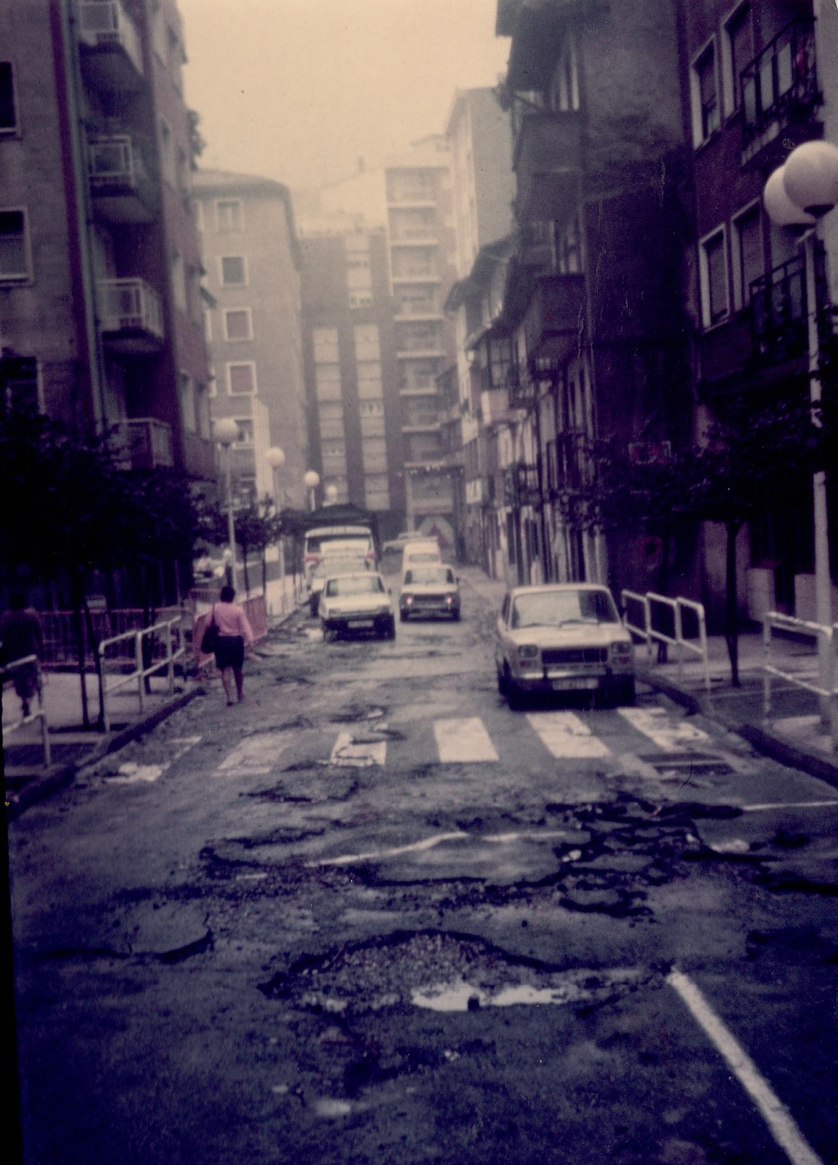 Se ve la el asfalto de la calle lleno de baches y roto por las inundaciones. Hay algunos coches aparcados y otros circulando.