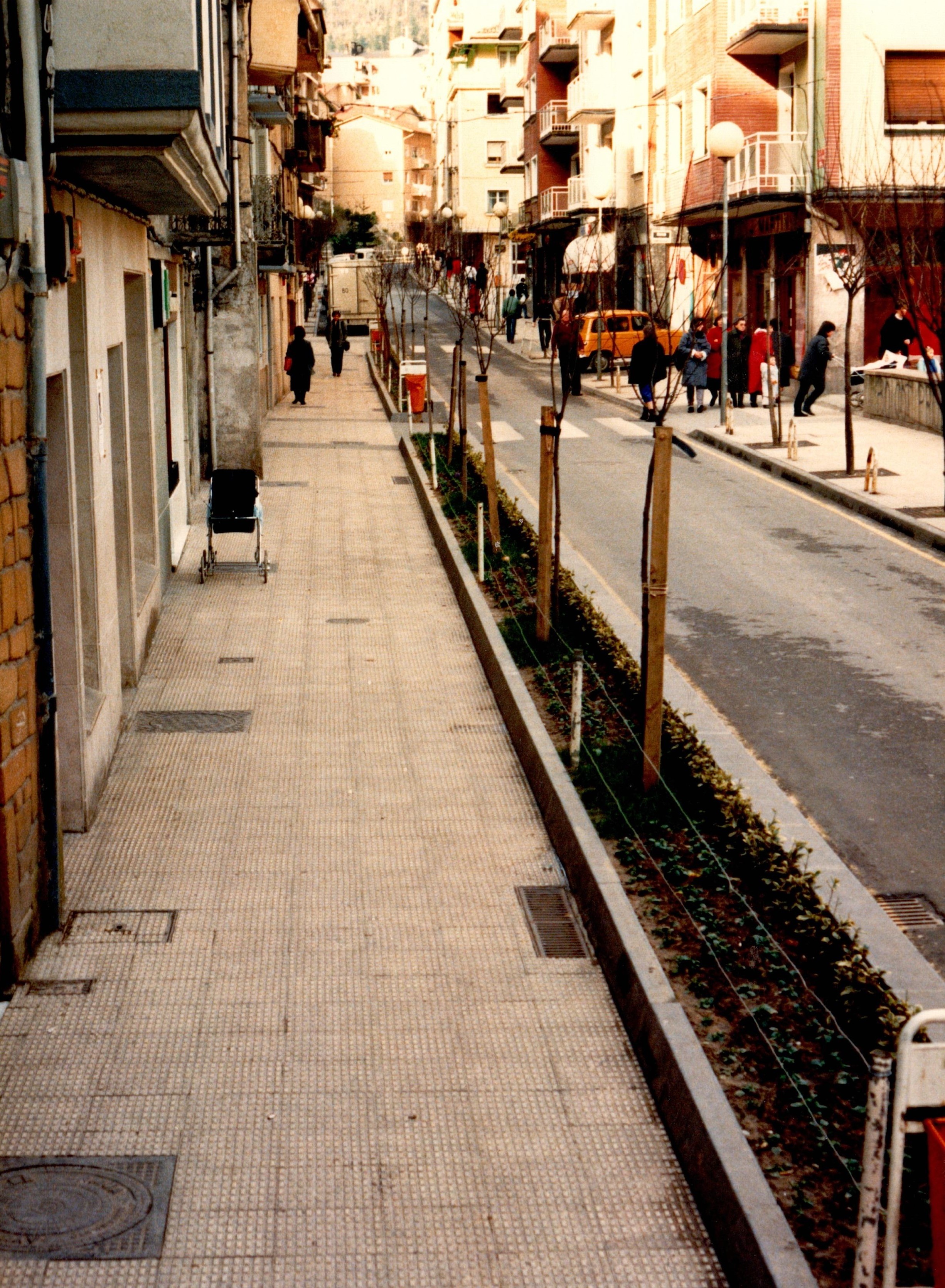 Se ve la acera y carretera de la calle Zubiaurre después de las obras de las tuberías. Hay un tramo nuevo de jardín muy estrecho y en vertical, paralelo a la carretera y separando la acera de la carretera. En ese pequeño jardín hay uno árboles recién plantados, muy finos.