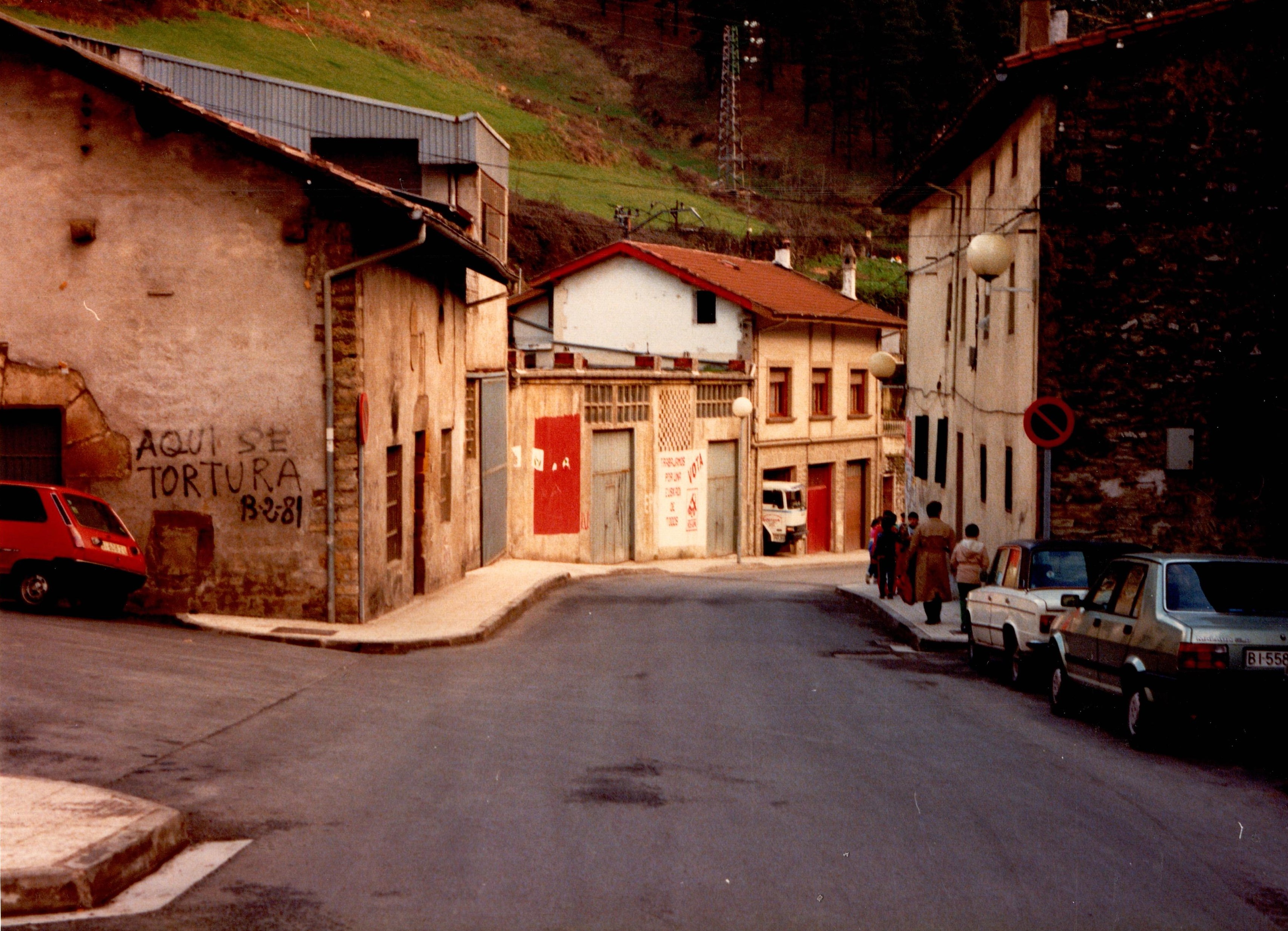 Fotografía tomada desde la carretera que atraviesa la calle Izelaieta. A la izquierda hay algunos edificios, al lado del primero en el lado izquierdo hay un coche rojo aparcado. En la pared se puede leer una pintada en negro en la que pone "aquí se tortura 13-2-1981. A la derecha hay algunos coches aparcados, se ven dos, uno gris y uno blanco. Por la acera derecha junto a las viviendas transitan algunos peatones.