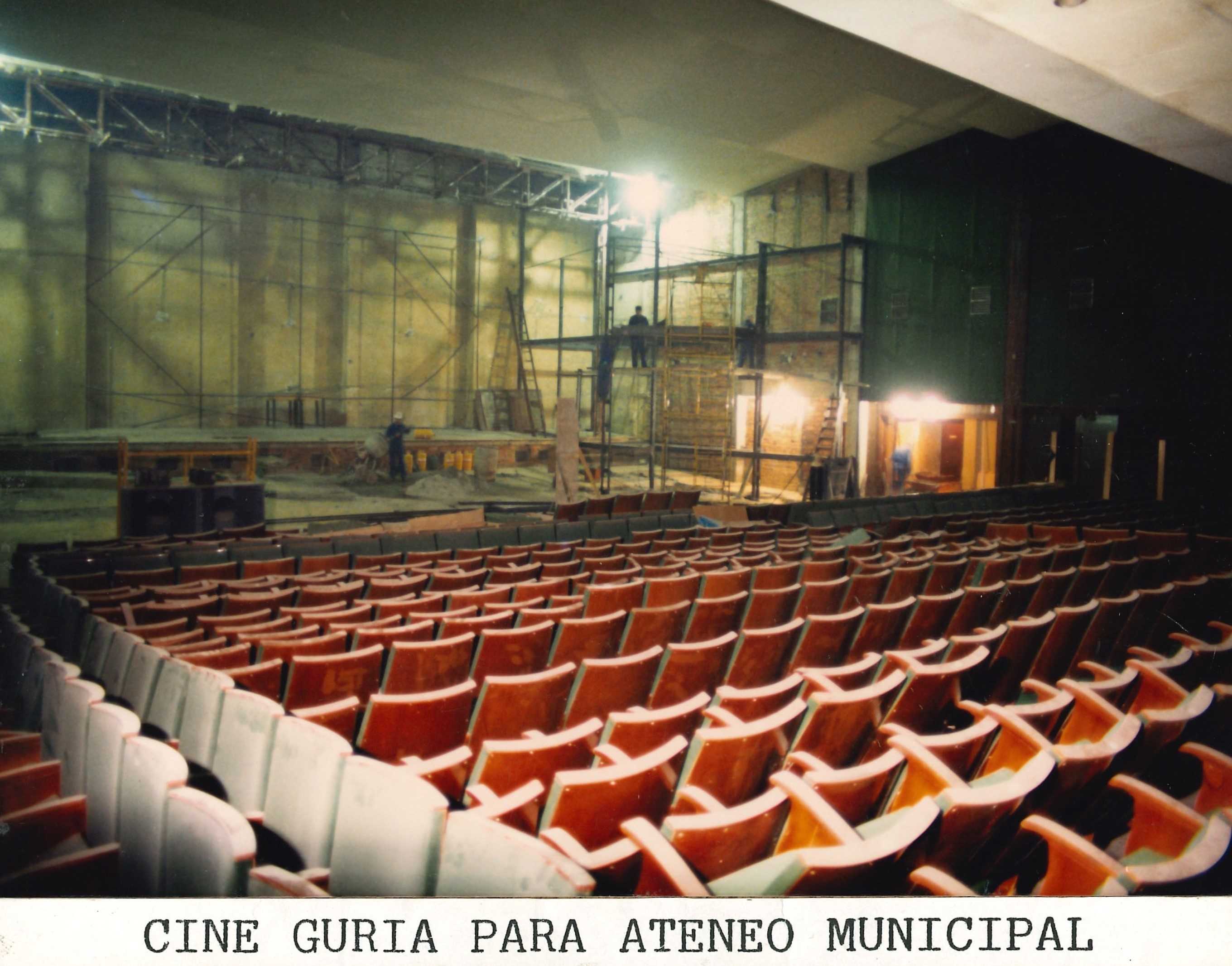 Se muestra el antiguo cine con las butacas, al fondo se ven unos andamios y la sala está iluminada con focos de obra. Se pueden ver algunos trabajadores en los andamios. Las butacas son rojas y verdes. 
