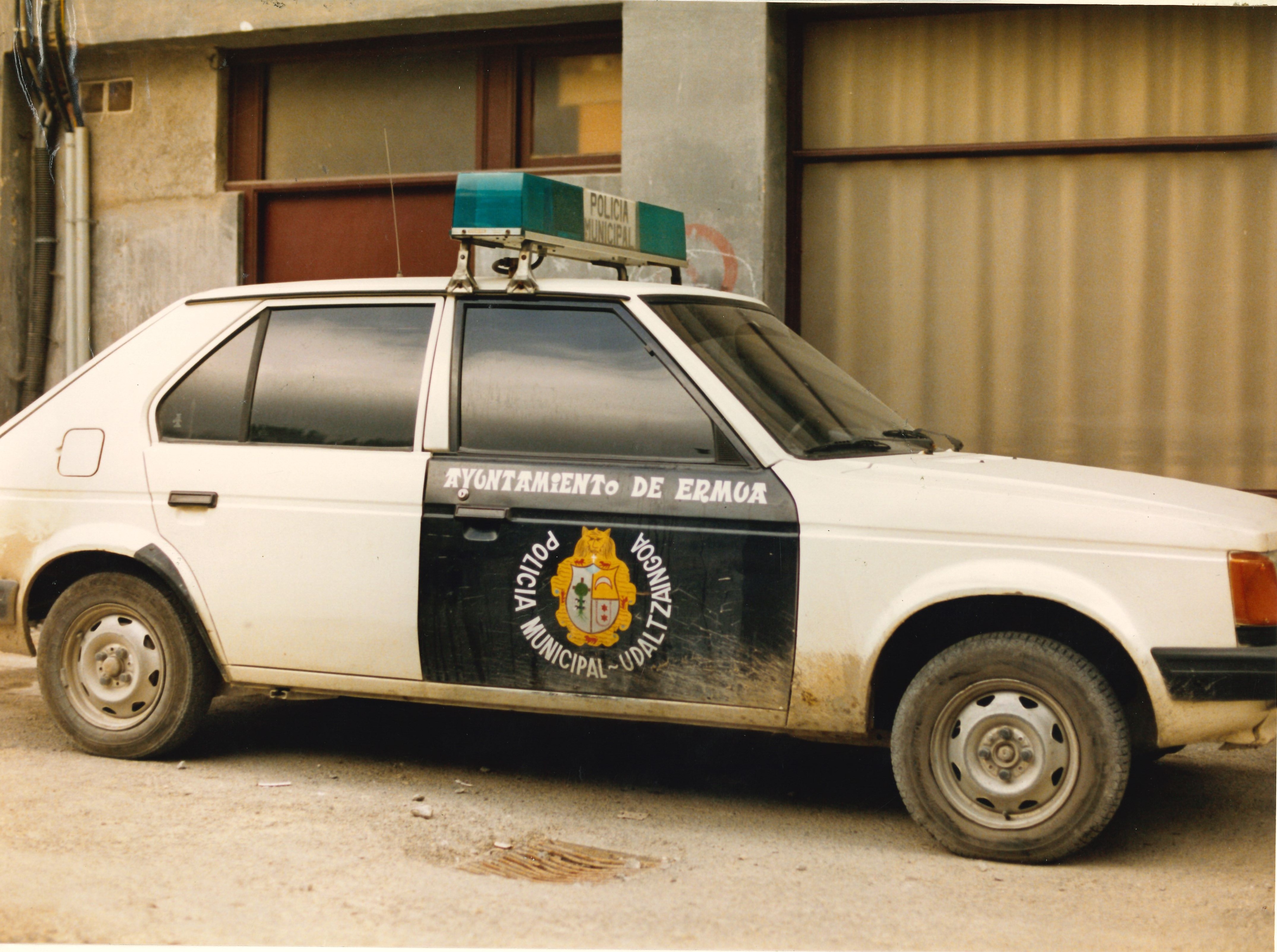 Se ve un vehículo (coche de cinco plazas). Solo se ve un lateral del coche, la parte del copiloto. En esa puerta, con fondo negro y letras blancas se puede leer: Ayuntamiento de Ermua y alrededor del escudo de la Villa de Ermua: POLICÍA MUNICIPAL - UDALTZAINGOA. 