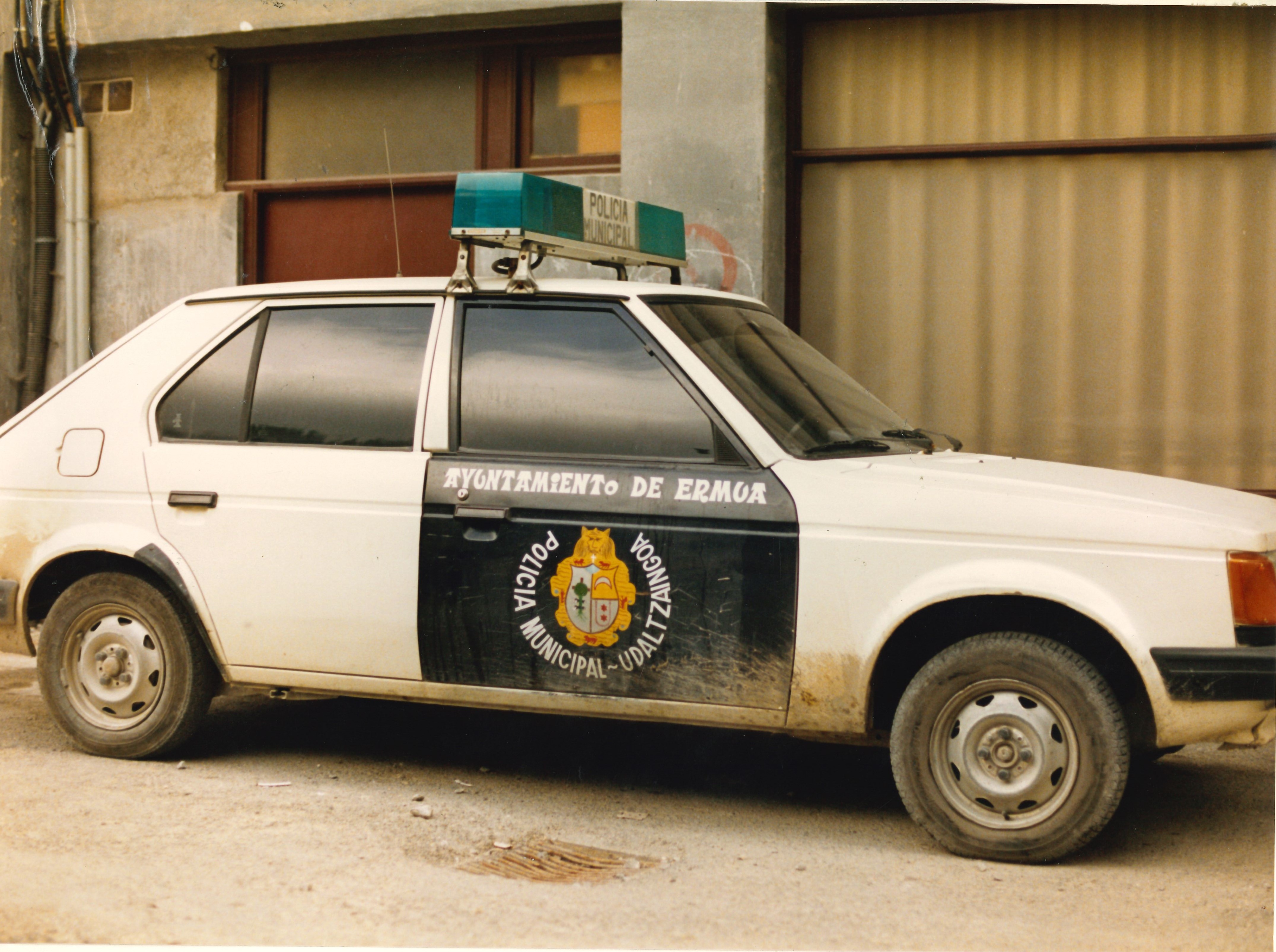 Se ve un vehículo (coche de cinco plazas). Solo se ve un lateral del coche, la parte del copiloto. En esa puerta, con fondo negro y letras blancas se puede leer: Ayuntamiento de Ermua y alrededor del escudo de la Villa de Ermua: POLICÍA MUNICIPAL - UDALTZAINGOA.