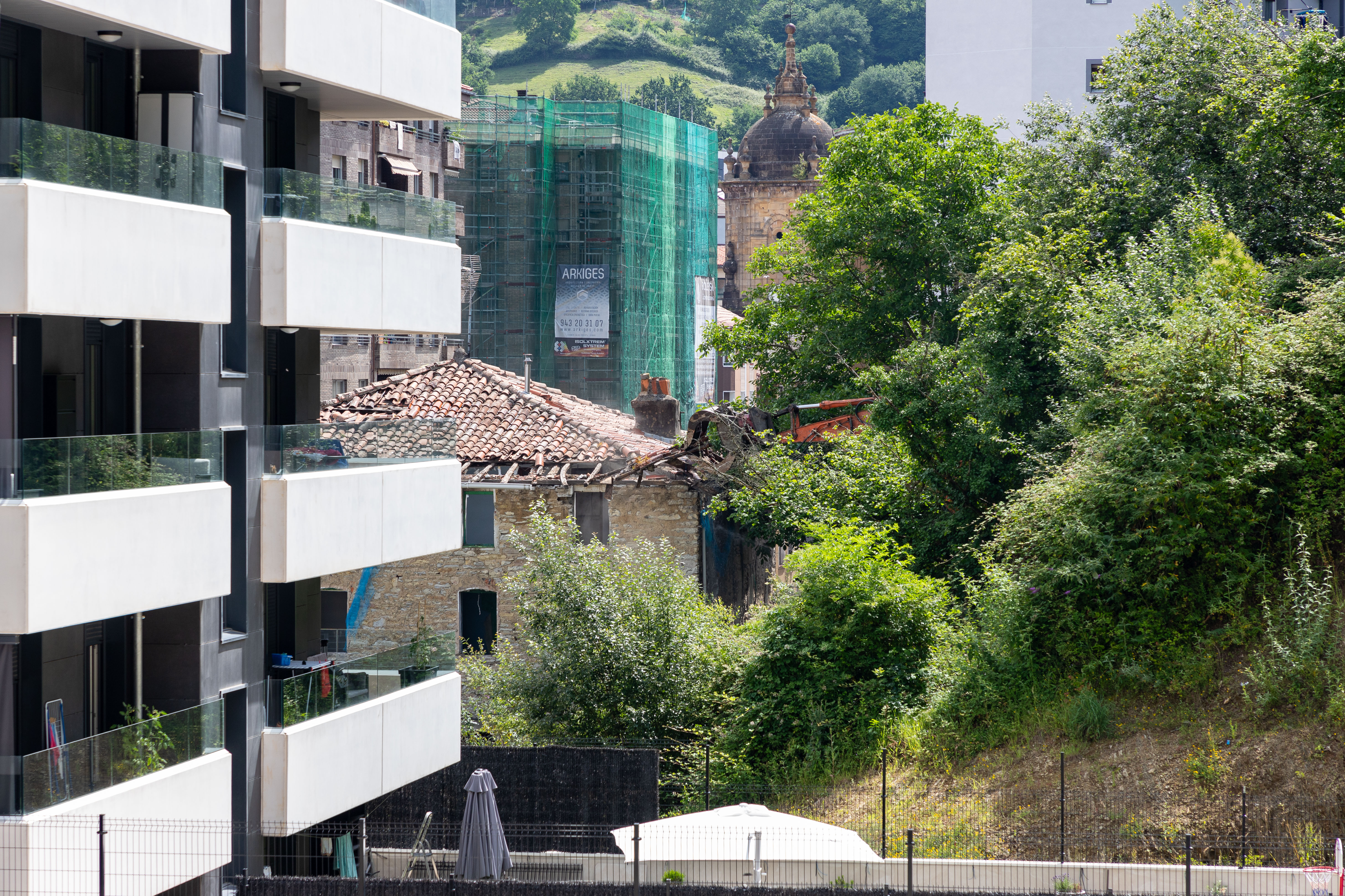 Es una fotografía en la que se ve a la izquierda un edificio nuevo con la fachada blanca y varios cristales en los balcones. A la izquierda se ven algunos árboles y arbustos verdes. En el centro pero con perspectiva más lejana el edificio que están derribando, un poco de la máquina que derriba el tejado y al fondo el centro urbano con una fachada que están restaurando y parte del edificio emblemático Marqués de Valdespina.