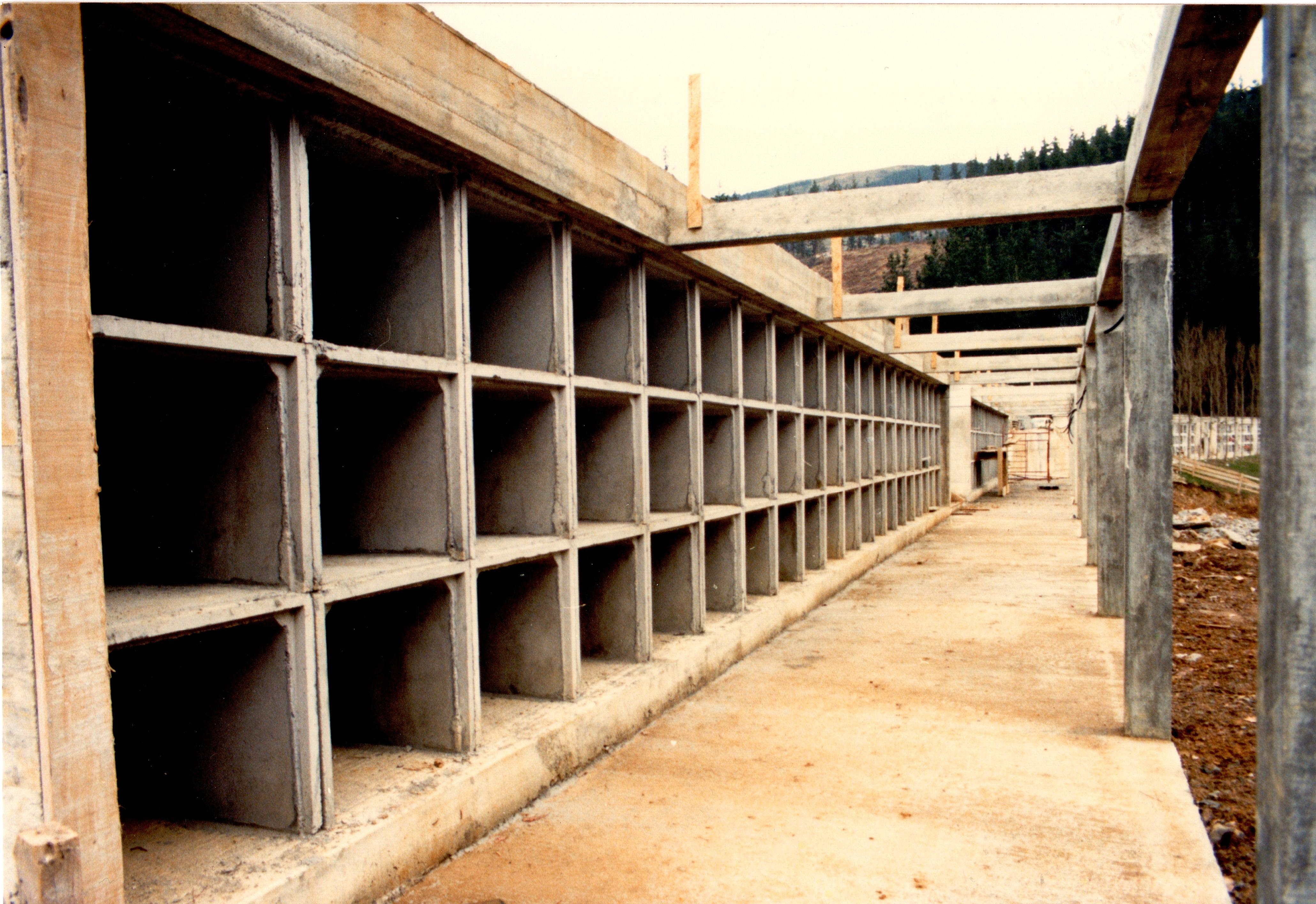 Se ven las estructuras recién hechas de lo que serán los nichos del cementerio. Tienen forma cuadrada y son grises, parecen hechos con cemento porque se ven los restos todavía. Delante de los nichos y encima de ellos hay una estructura de barrotes y un pasillo de piedra para caminar delante de ellos.