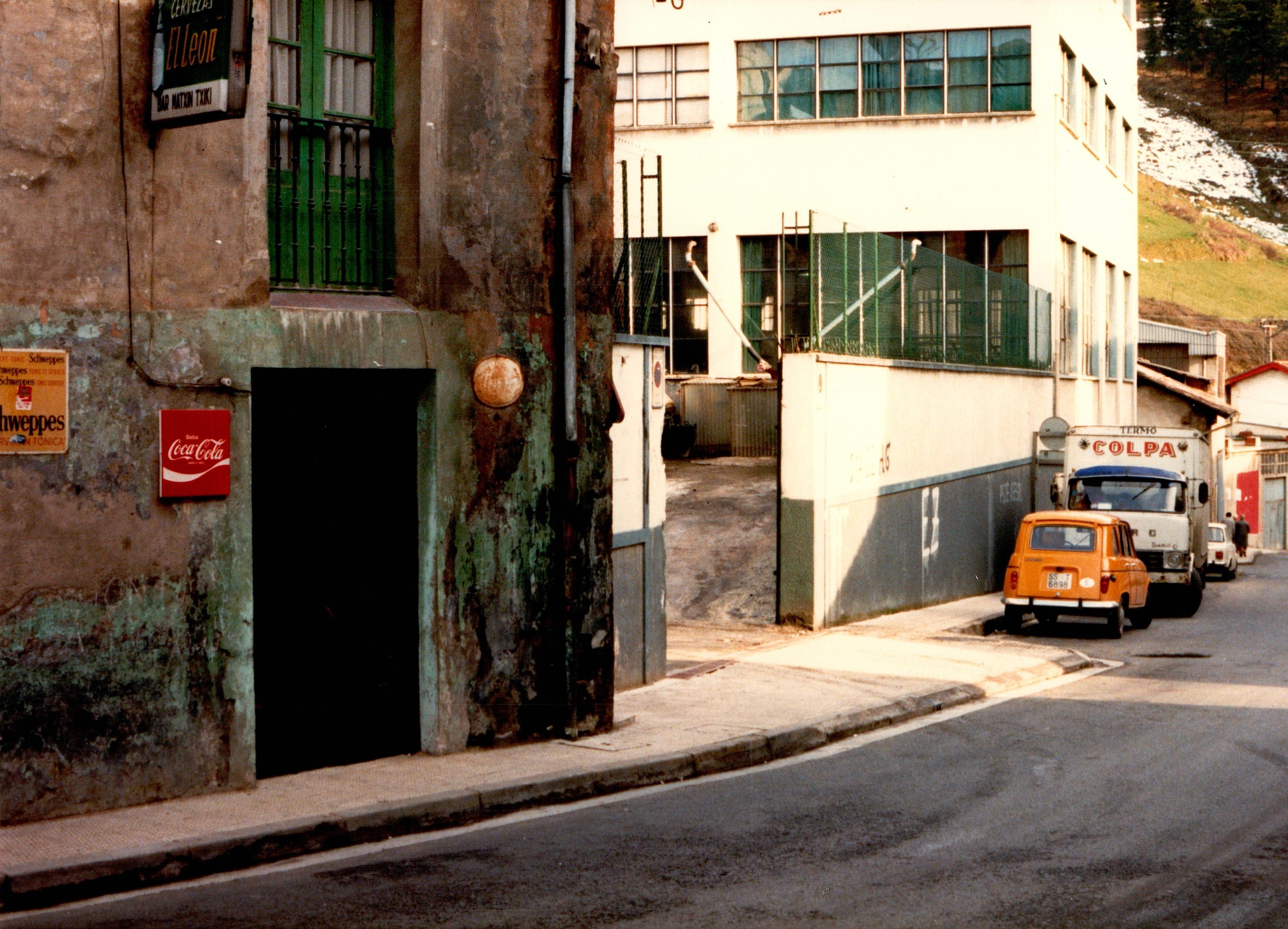 Se muestra la parte izquierda de la calle Izelaieta, lo primero que se ve es un bar, tal y como indica un cartel "Bar Matxin Txiki". También tiene un cartel rojo con letras blancas que pone "CocaCola" y otro amarillo en el que pone Schweppes. El edificio siguiente es la fábrica "Cuchillería del Norte", es un edificio blanco con ventanas y puertas de acceso. A sus pies, en la carretera hay coches aparcados en paralelo, el primero en verse un coche color amarillo-naranja. Después de ese coche hay un camión.