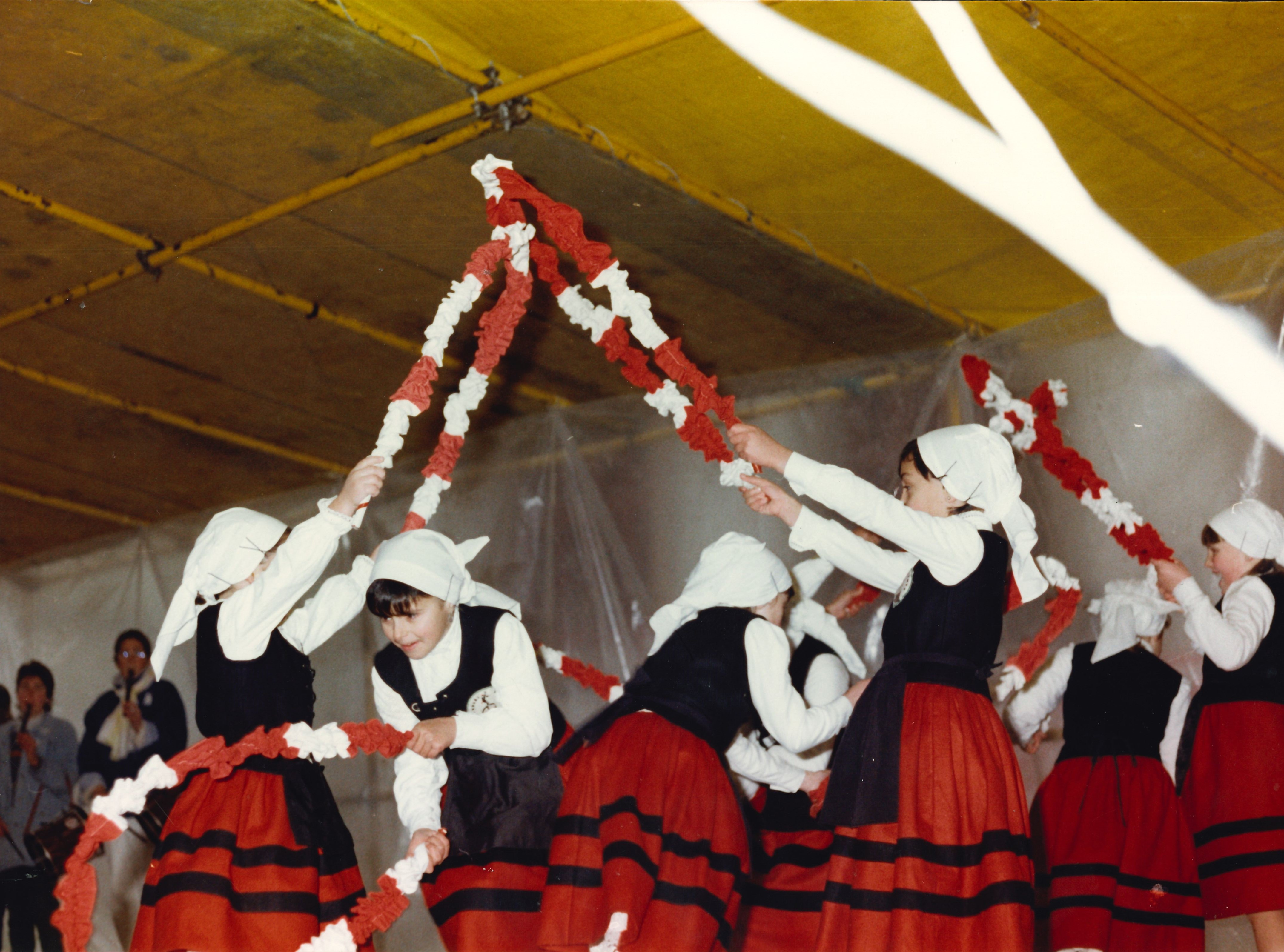 Un grupo de niñas está bailando con arcos de flores blancos y rojos. Visten con el traje tradicional de las dantzas, en este caso falda roja con dos rayas horizontales negras. También llevan pañuelo blanco en la cabeza.