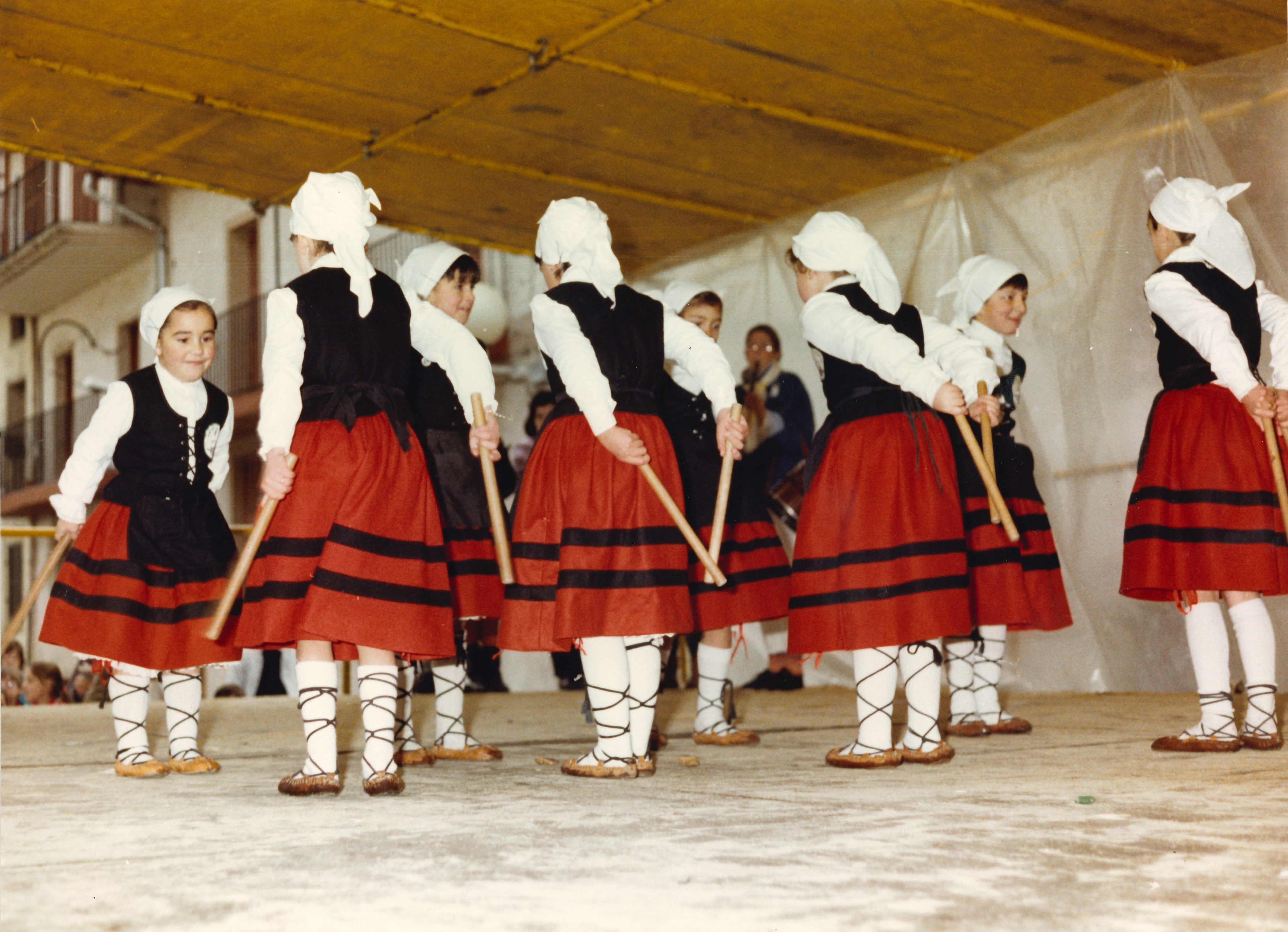 Un grupo de niñas bailan con makilas (palos de danzadoras) en un escenario. Visten el traje típico de dantzas vascas, alpargatas marrones, falda hasta las rodillas roja con dos rayas horizontales negras, pañuelo blanco en la cabeza y camisa blanca.