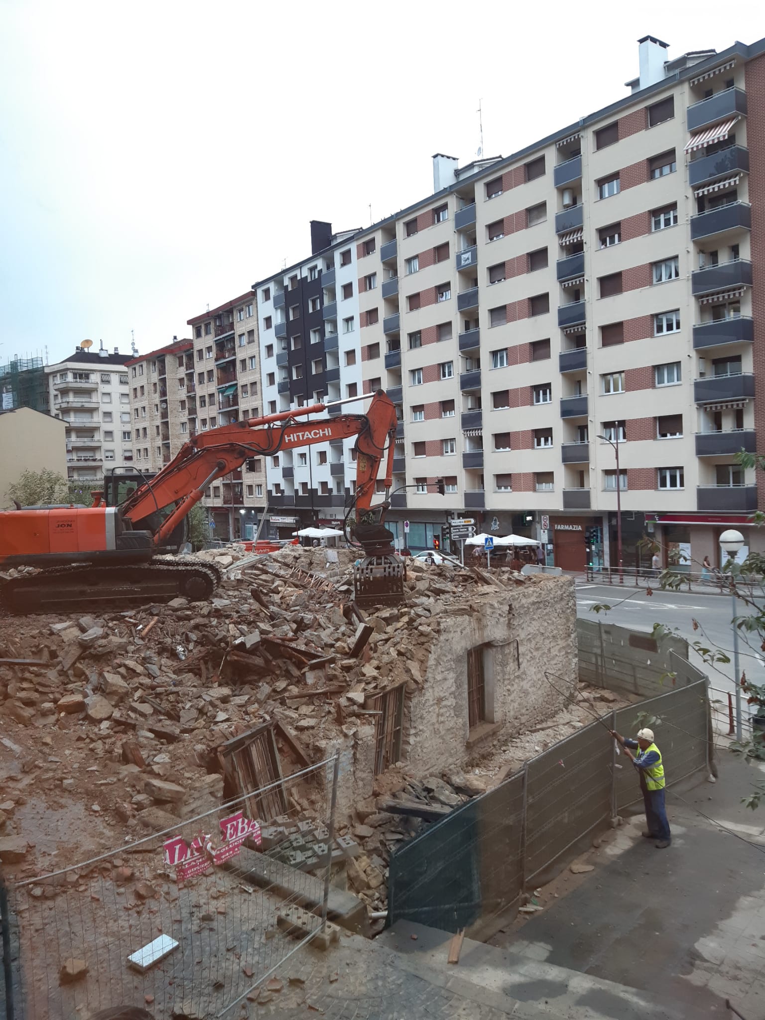 Una excavadora naranja color butano está en acción derribando desde el interior de la estructura del edificio o lo que queda de ella. Todo el edificio está derribando menos las paredes de la planta baja, hay polvo que se levanta al caer las piedras y mover los escombros. En el suelo hay muchos escombros, la mayoría de las ventanas están a punto de caerse y el entorno está vallado con vallas metálicas y otras metálicas también pero con malla verde.