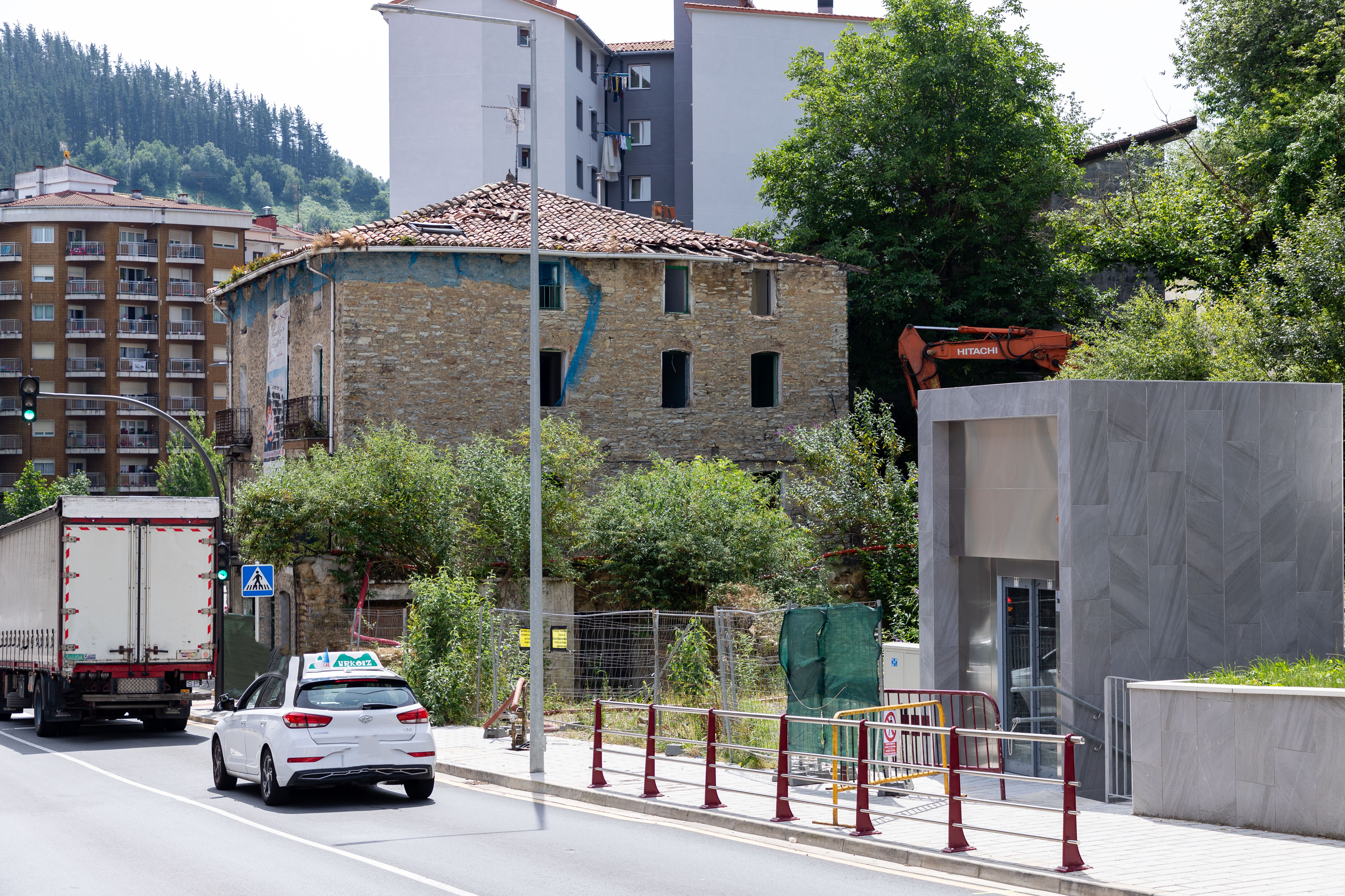 Fotografía tomada desde la carretera donde se ve el edificio que van a derribar. Se ve a la izquierda un ascensor de las nuevas viviendas construidas cerca de la casa que van a derribar. Alrededor de la vivienda del derribo está delimitada la zona de forma que los peatones no pueden transitar. Se ve un coche de autoescuela blanco y delante un camión circulando por la carretera. También pueden observarse dos edificios detrás del edificio que van a derribar.