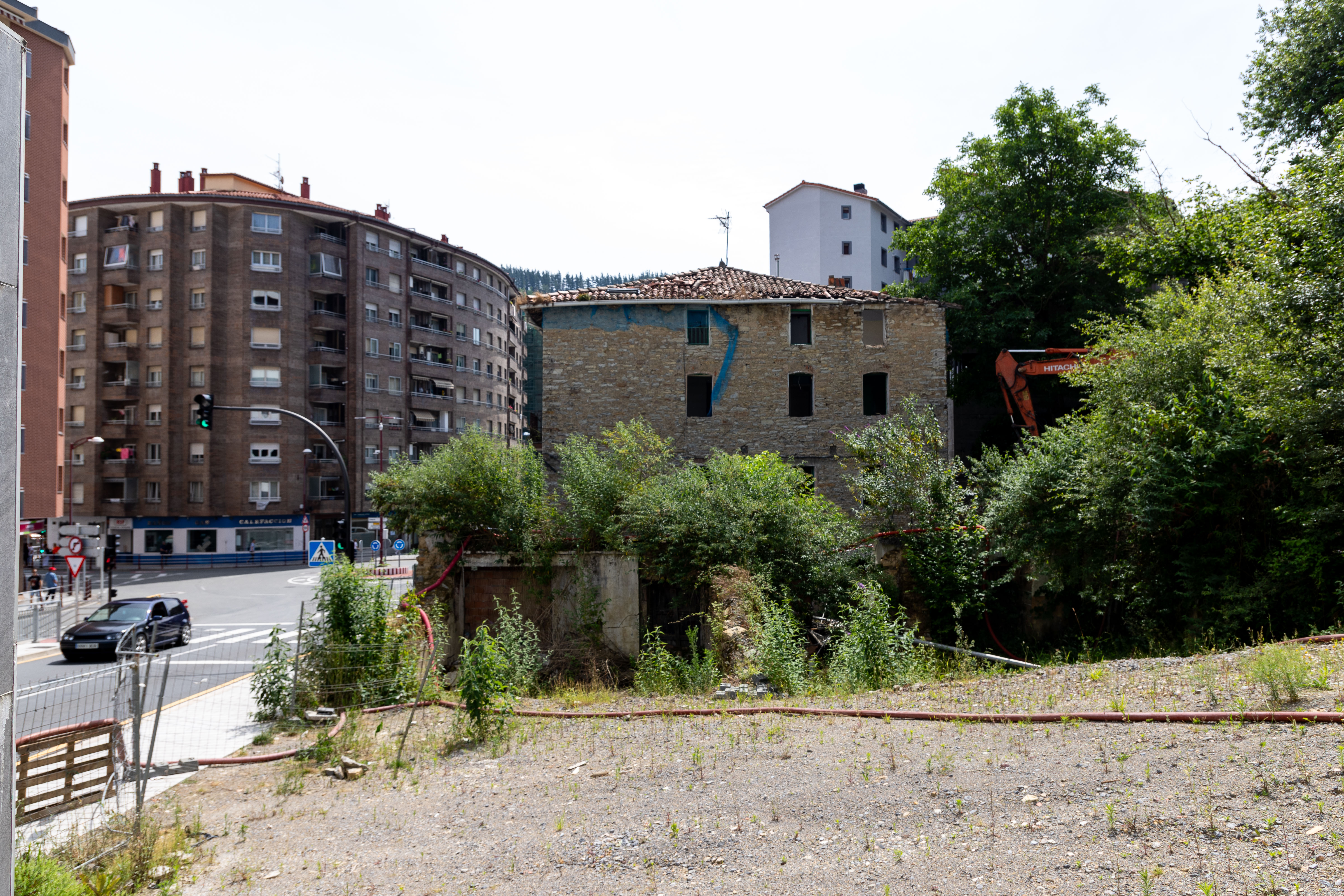 Fotografía que muestra un lateral de la fachada del edificio que van a derribar. Se ve una parcela vacía, árboles, arbustos y parte de la carretera. Algunos edificios cercanos al edificio en cuestión se muestran también. A la derecha, entre los árboles y la vegetación