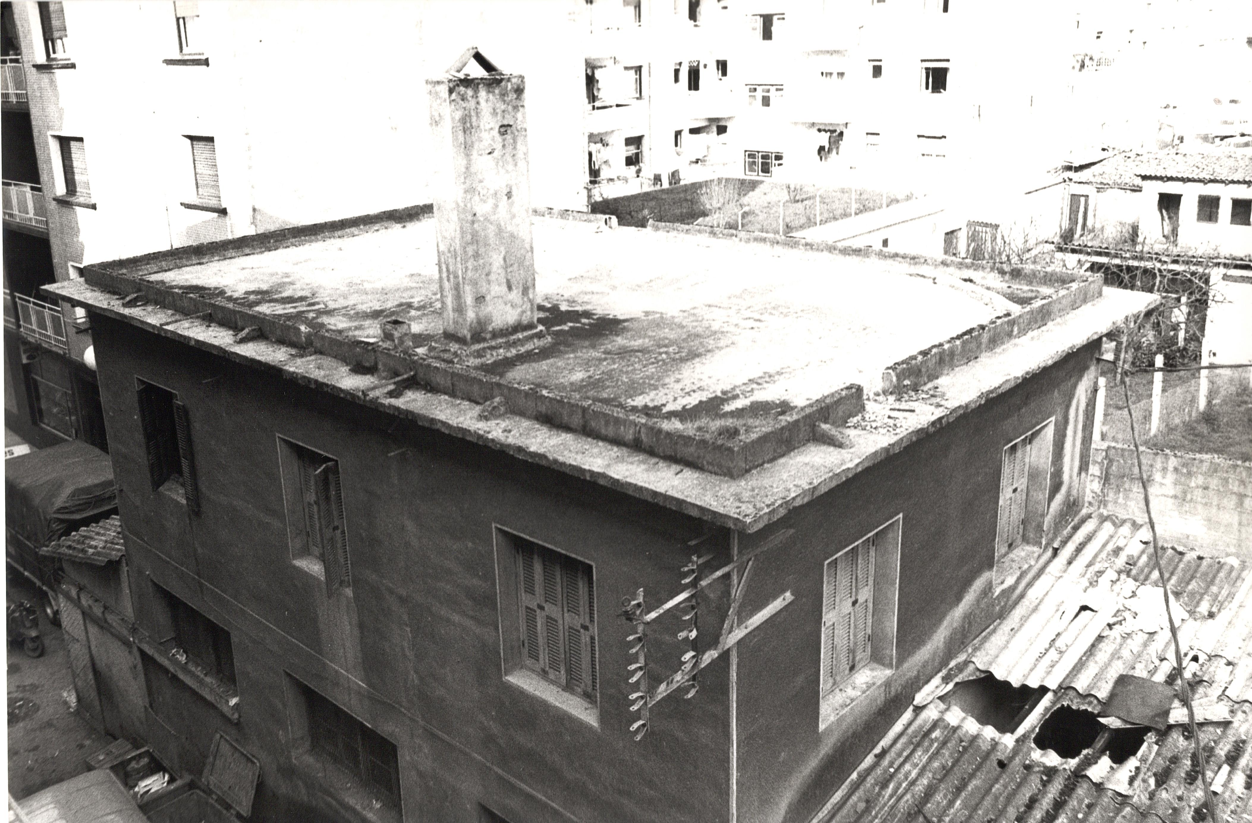 Se ve un edificio con una chimenea en el tejado, una chimenea de piedra. Cerca hay otros edificios pero que no se ven con nitidez por la calidad de la fotografía, Es una foto en blanco y negro. Hay varias ventana y el edificio tiene dos plantas. 