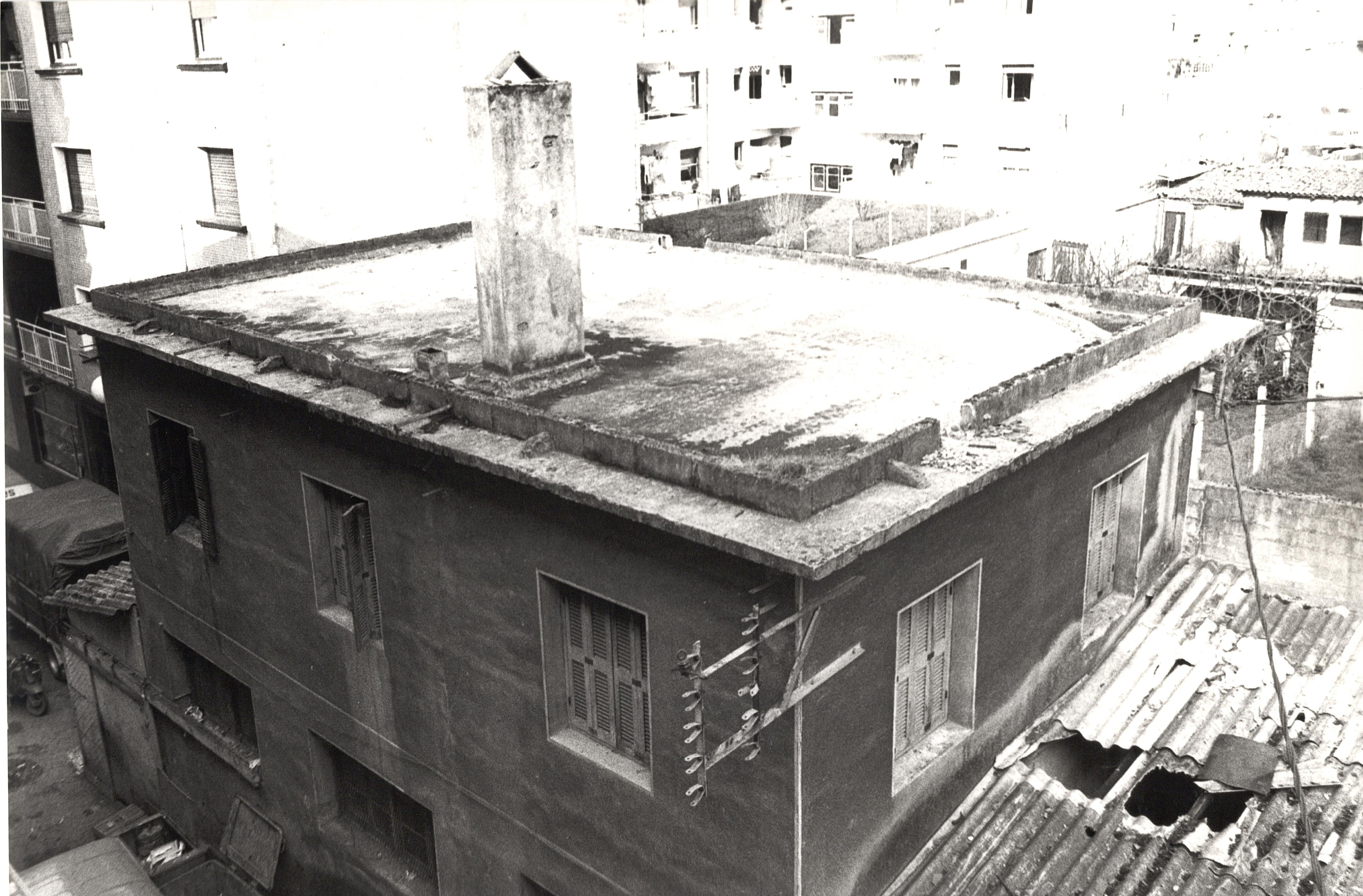 Se ve un edificio con una chimenea en el tejado, una chimenea de piedra. Cerca hay otros edificios pero que no se ven con nitidez por la calidad de la fotografía, Es una foto en blanco y negro. Hay varias ventana y el edificio tiene dos plantas.