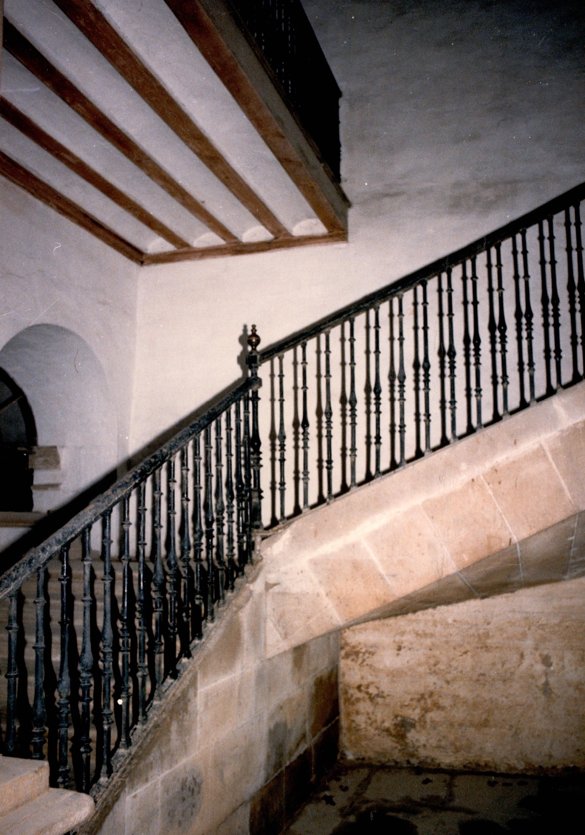 Se muestra una escalera con barandilla metálica negra. El resto de la estructura es de piedra, tanto los suelos, como las paredes y las escaleras que conducen a las plantas superiores. Se ven dos tramos de escaleras que se intuyen gracias a las barandillas. En la parte superior hay algunos elementos de madera pero predomina la piedra de color arena cálida. Las paredes están pintadas de blanco.