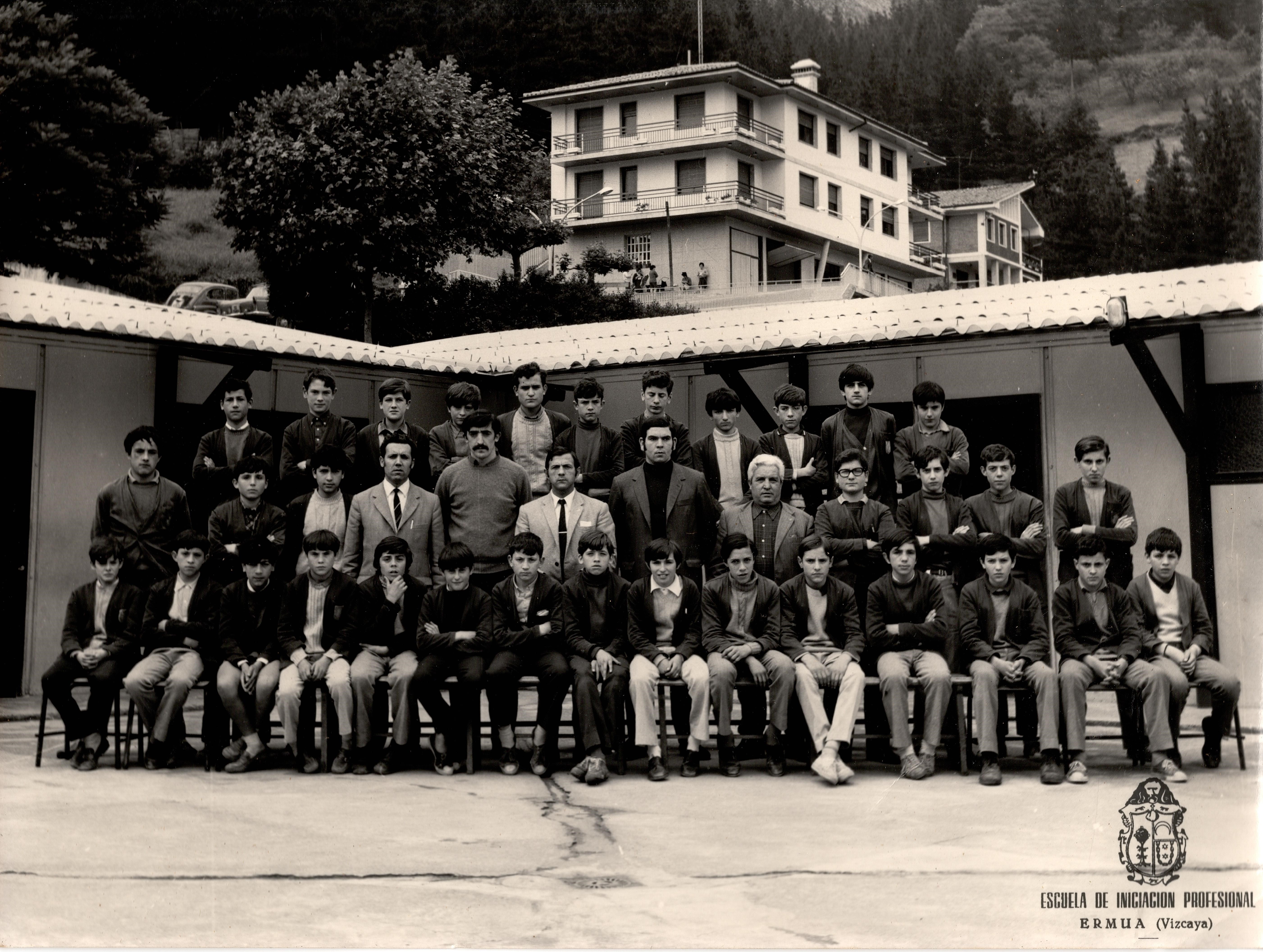 Tres filas de jóvenes están posando, son alumnos y profesores de la escuela de iniciación profesional de Ermua. En la primera fila hay 15 chicos sentados, en la segunda fila hay siete alumnos y cinco profesores/adultos en pie y en la última fila hay once alumnos en pie. La fotografía es en blanco y negro. Detrás de ellos se ven algunas paredes, el tejado de las instalaciones donde recibían la formación, y fuera, más atrás se ven dos coches aparcados y un par de viviendas.