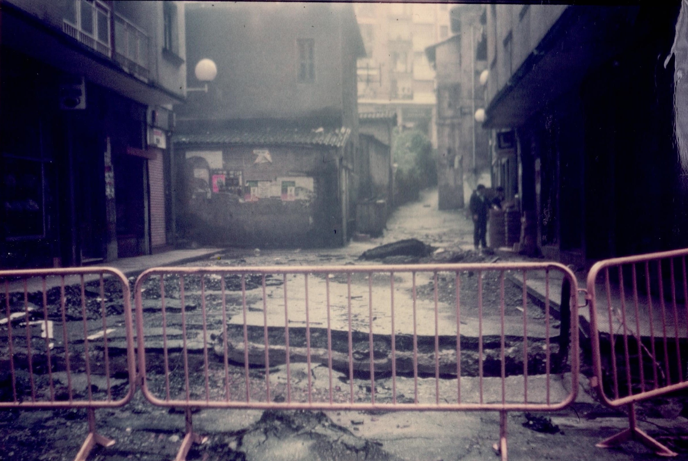Se ven tres vallas amarillas que impiden el paso a la calle Zubiaurre. Detrás de las vallas está todo el asfalto levantado, con grietas y en malas condiciones debido a las consecuencia de unas inundaciones devastadoras. A los lados hay edificios y al fondo hay dos personas en lo que parece la parte de fuera de un bar.