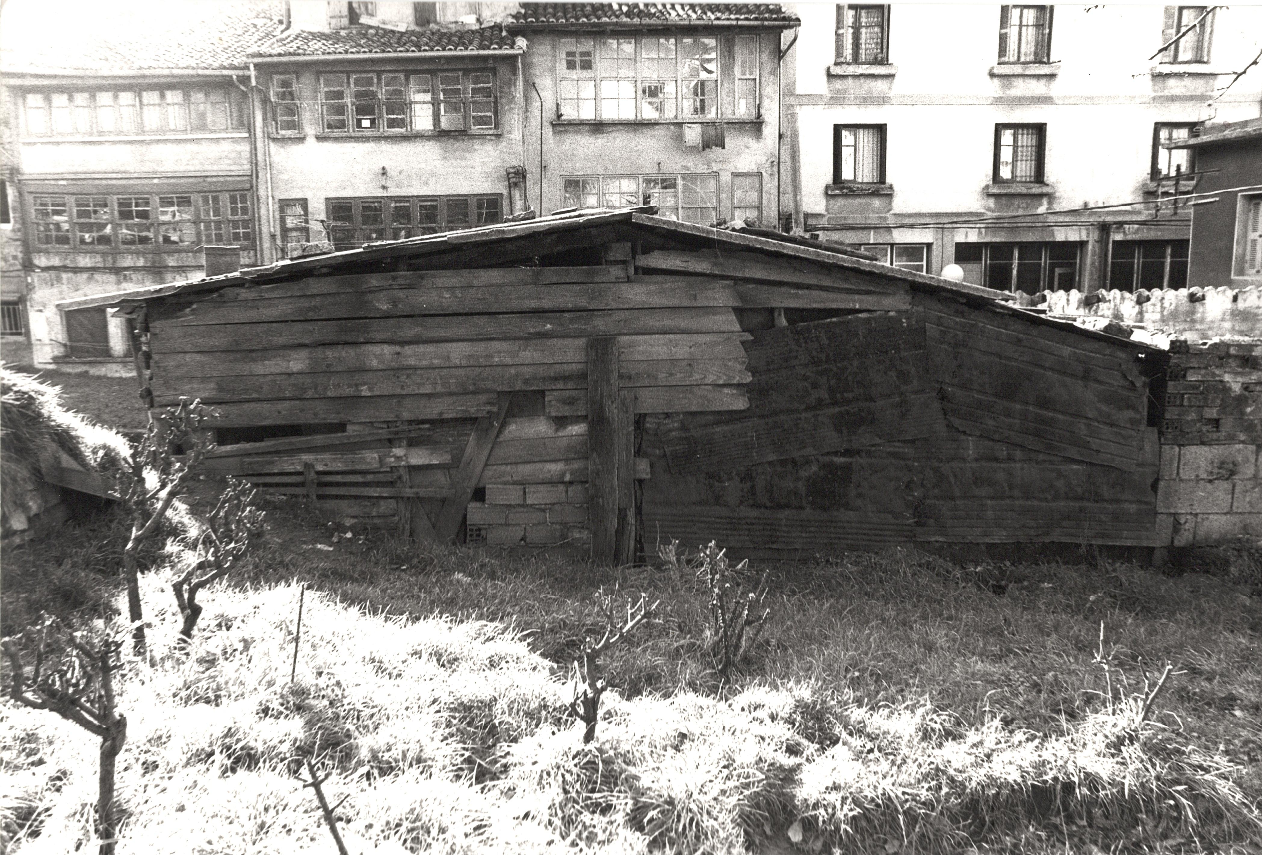 Se ve la trasera de una pequeña edificación de madera, una caseta más bién. Son tablas de madera y un tejado no muy robusto. Tendrá una altura máxima de dos/tres metros. Al fondo se ven cuatro edificios, los tres primeros empezando por la izquierda son viviendas. 