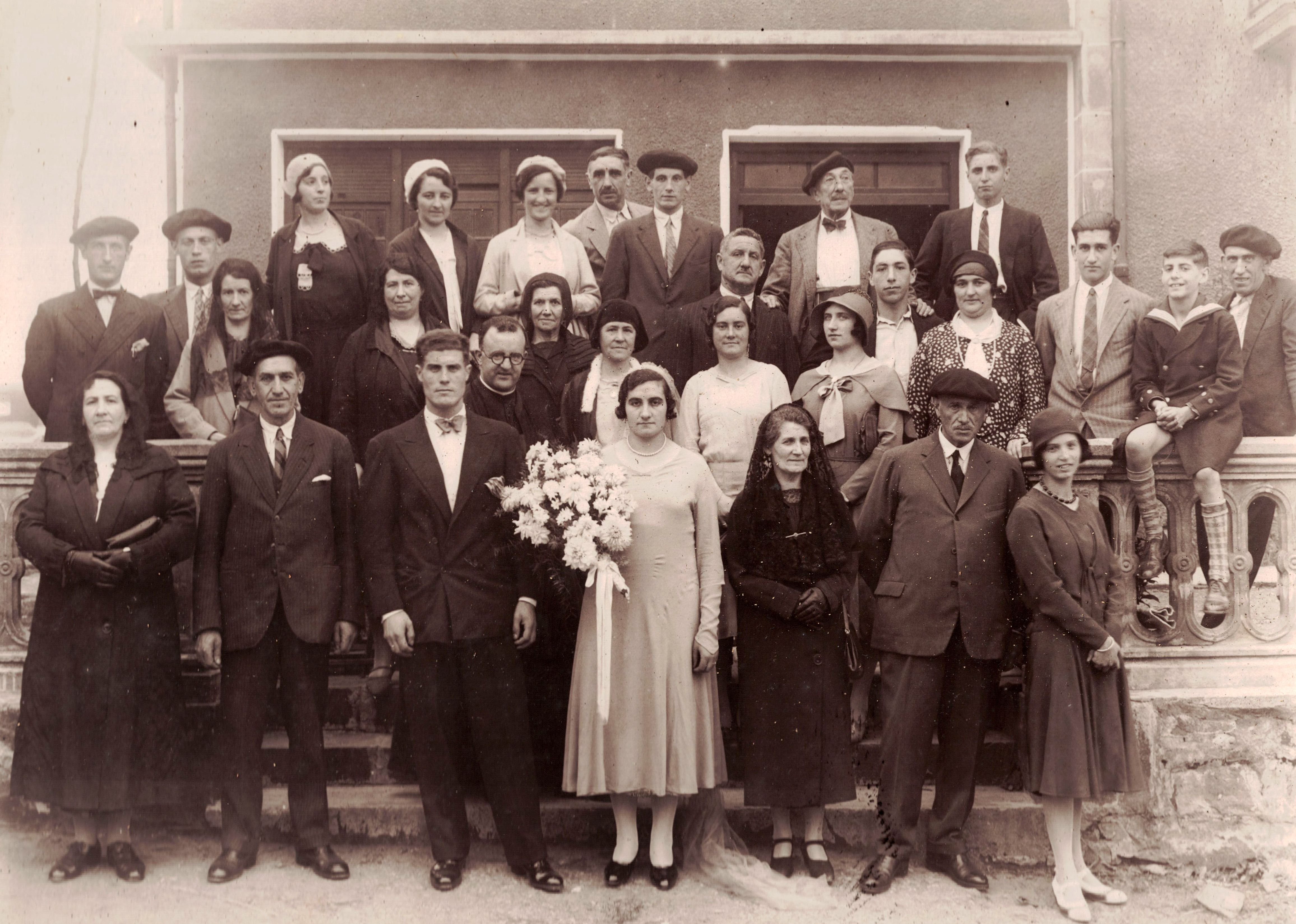 Fotografía de las invitadas e invitados y los recién casados el día de su boda. Muestra 14 mujeres y 14 hombres. En primera línea central están Juana Solozabal sujetando un ramo de flores y Félix Izaguirre a su lado vestido de traje. La fotografía es en blanco y negro.