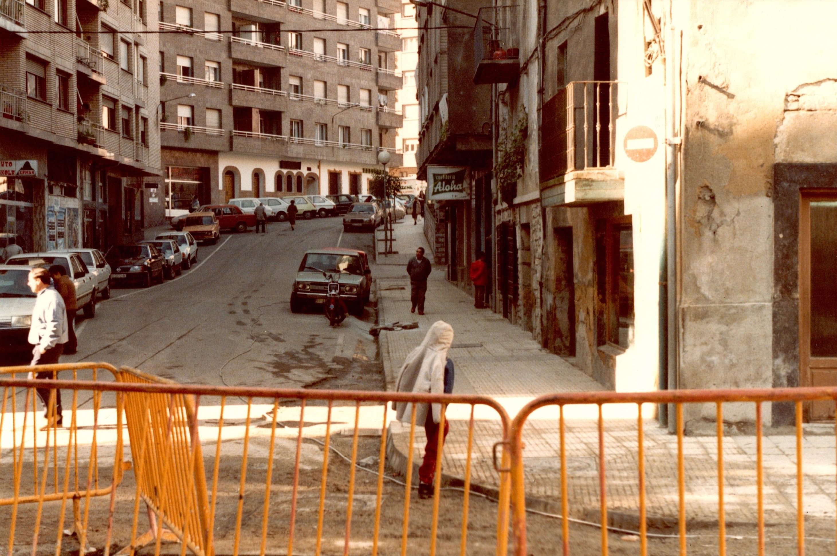 Se ven cinco vallas amarillas de obra separando la calle Zubiaurre y la calle Probaleku. La calle está sombría porque el sol no entra debido a los edificios. Hay edificios a la izquierda y a la derecha. También hay varios coches aparcados a las orillas de la carretera en paralelo, más coches a la izquierda que a la derecha. En la fotografía aparecen diez personas aproximadamente caminando en diferentes puntos de esta calle.