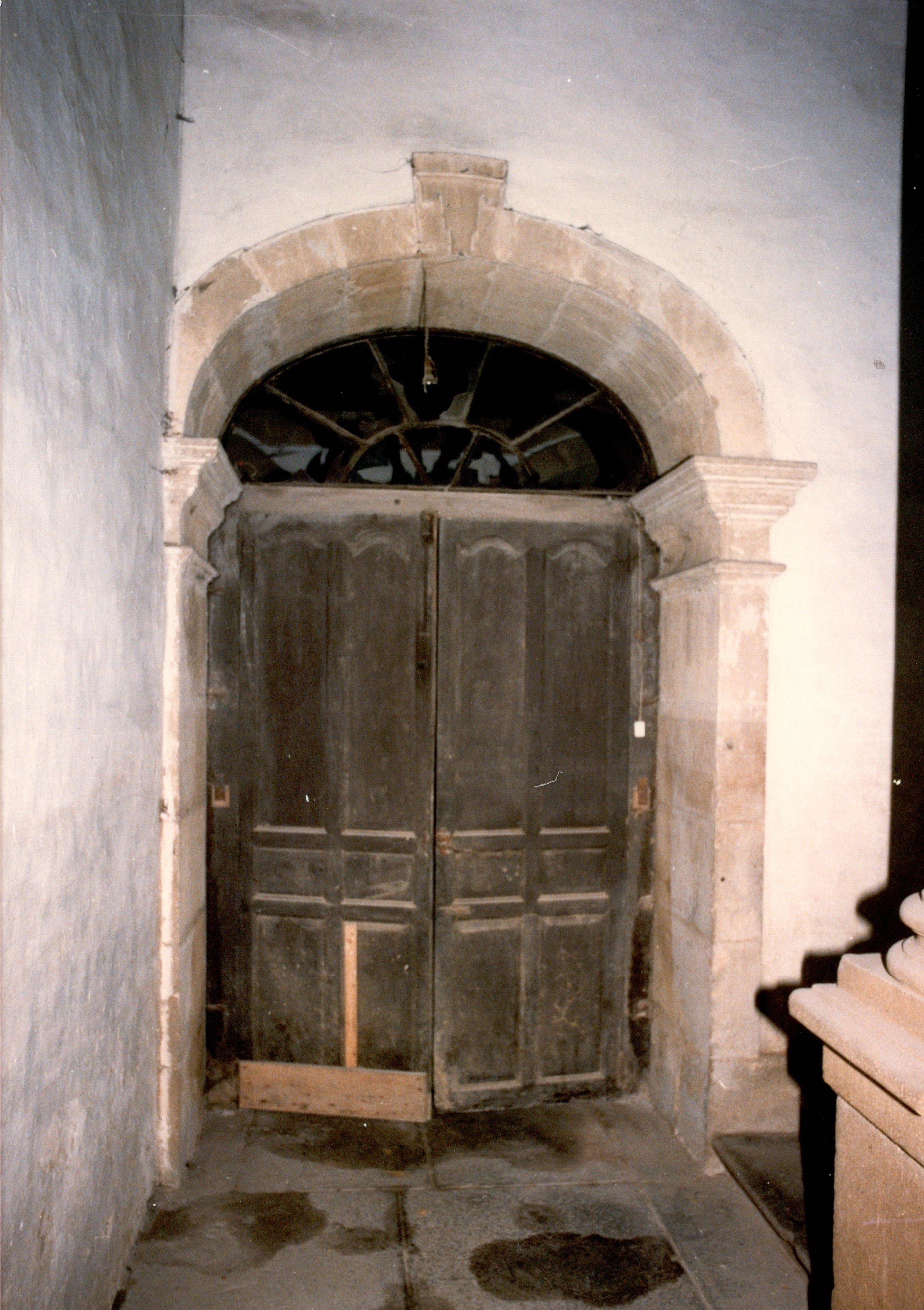 Se ve una puerta de madera, amplia, de doble hoja en condiciones regulares. Se ve que la madera es vieja, no brilla pero hace su función. La fotografía está sacada con flash, esa es la luz que recibe la puerta. A la izquierda hay una pared blanca y a los lados de las puertas hay elementos decorativos de columnas y un arco como elemento arquitectónico. El suelo tiene varias manchas, es de piedra pero más oscura.