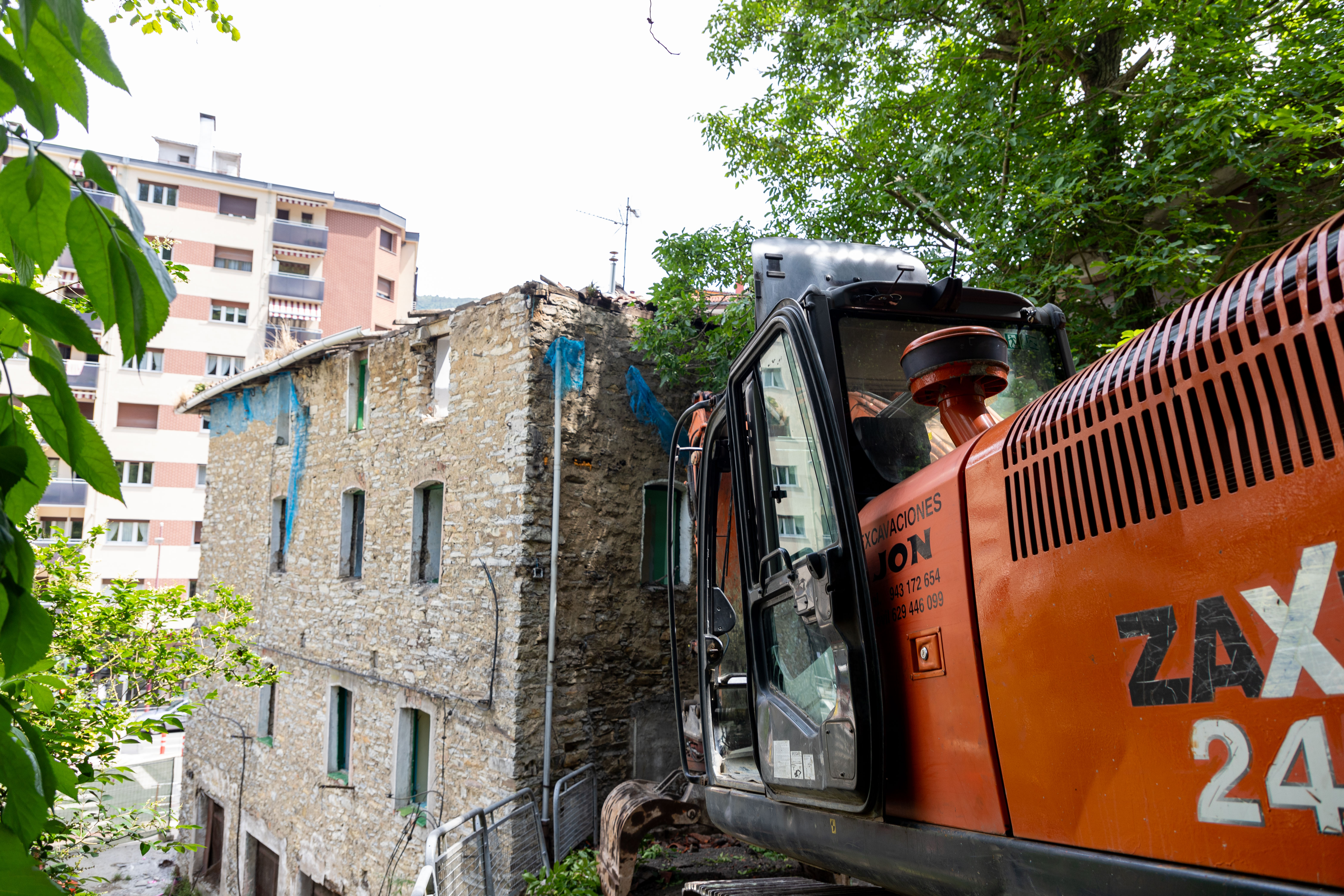 Se ve una máquina de excavación naranja trabajando en el derribo del edificio. En ella se puede leer "Excavaciones Jon" y dos números de teléfono: 943172654 y 629446099. Se está trabajando en el tejado y se ve la parte trasera y lateral del edificio. Hay varias vallas delimitando la zona y de fondo hay un edificio. Encima y a la derecha de la máquina hay un árbol verde.