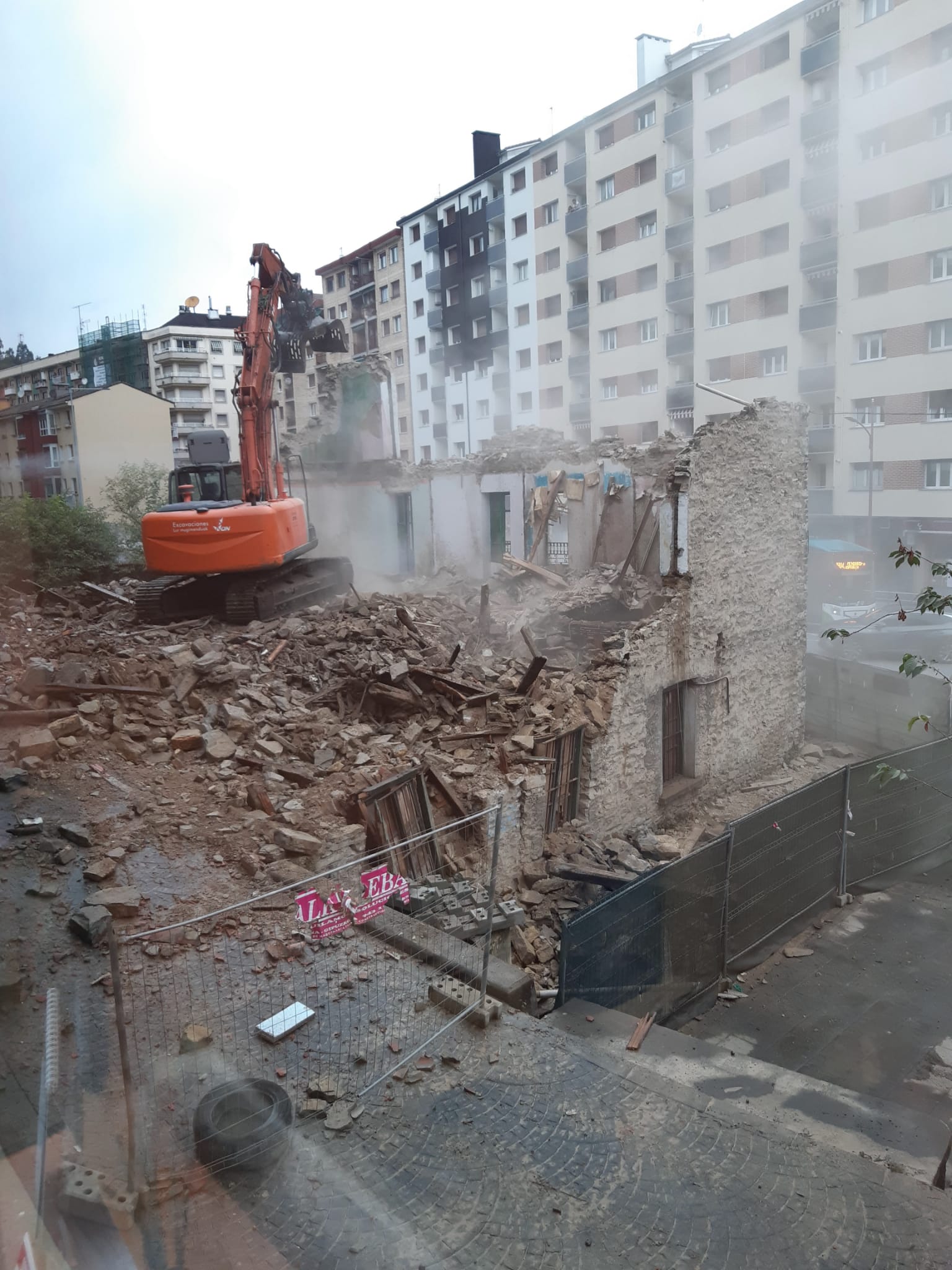 Una excavadora naranja color butano está en acción derribando desde el interior de la estructura del edificio o lo que queda de ella. Más de la mitad del edificio está derribado, hay polvo que se levanta al caer las piedras y mover los escombros. También hay agua que parece provenir de una manguera. En el suelo hay muchos escombros, la mayoría de las ventanas están a punto de caerse y el entorno está vallado con vallas metálicas y otras metálicas también pero con malla verde.