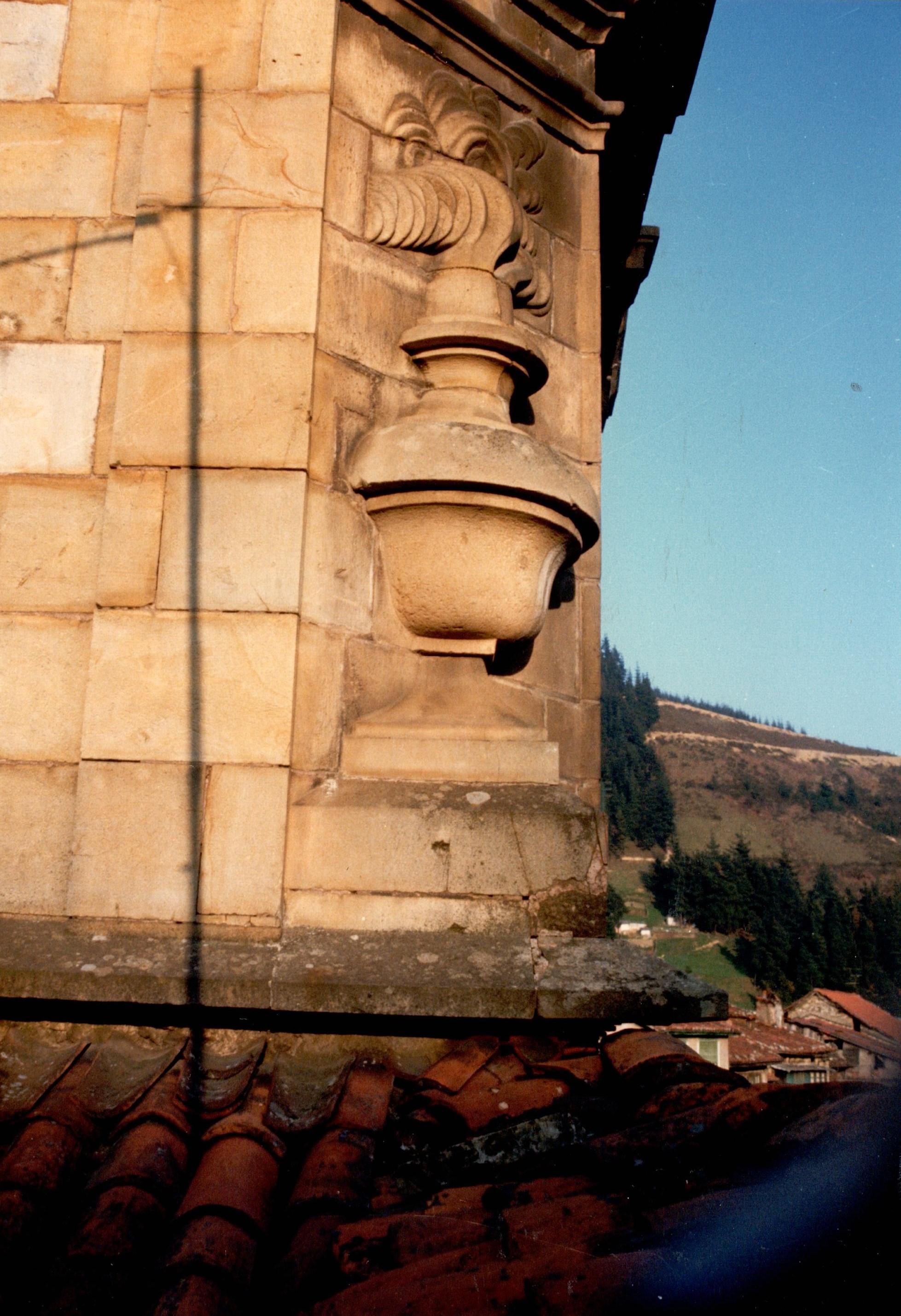 Se ve parte del tejado del edificio, de ladrillos rojos. Se fotografía principalmente la estructura que sostiene la cúpula del Palacio. El color de esa estructura de piedra es arena tostada, clarita. Al fondo se ven algunas casas, una parcela de hierba verde, árboles y monte. El cielo está azul claro, muy despejado.
