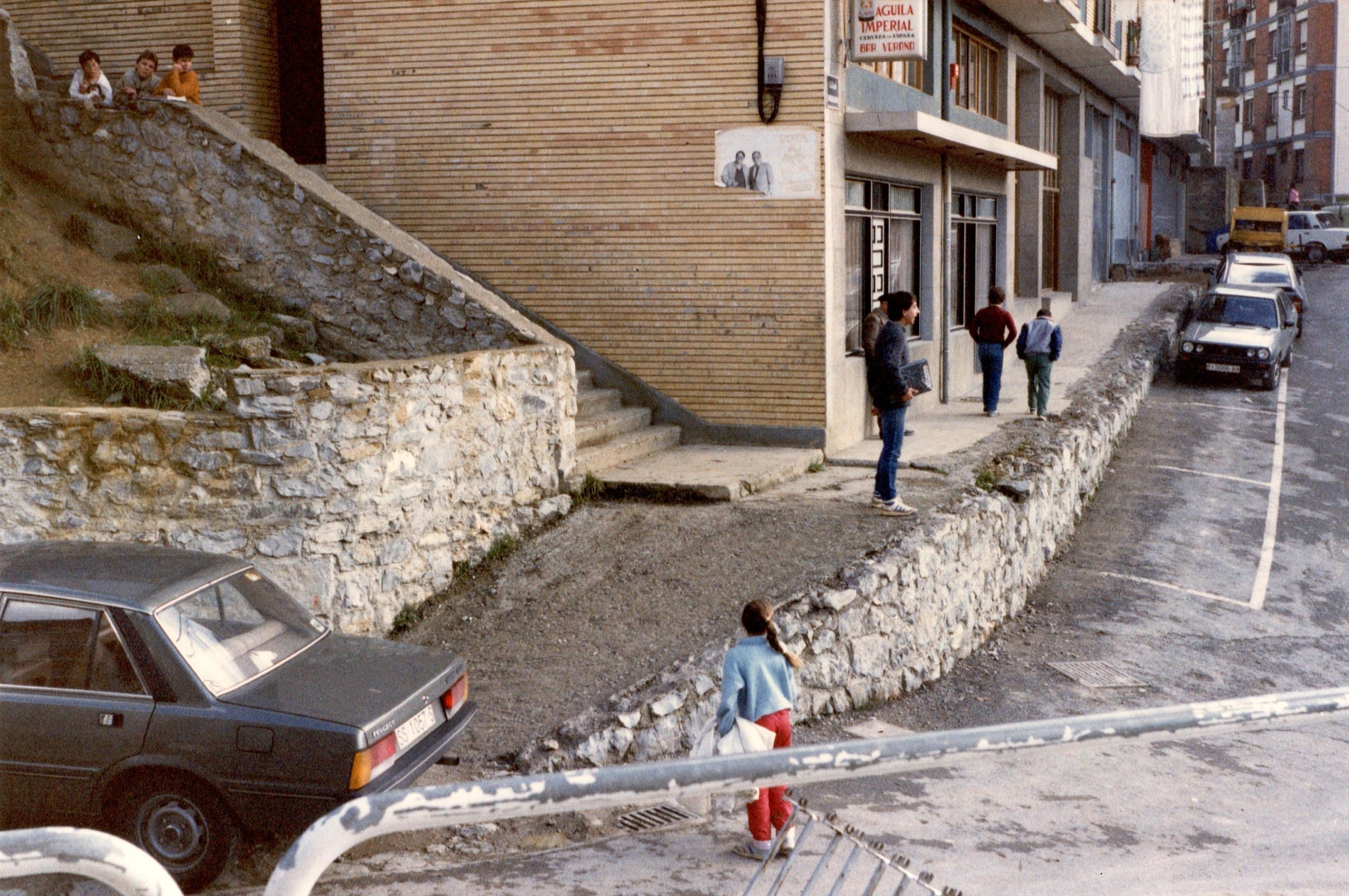 Se muestran las obras, algunos muros nuevos, se ve cementoreciente entre unas escaleras y la carretera. Hay un coche gris a la izquierda aparcado y más alante otros en paralelo al arcén. En las escaleras hay tres niños mirando asomados. 