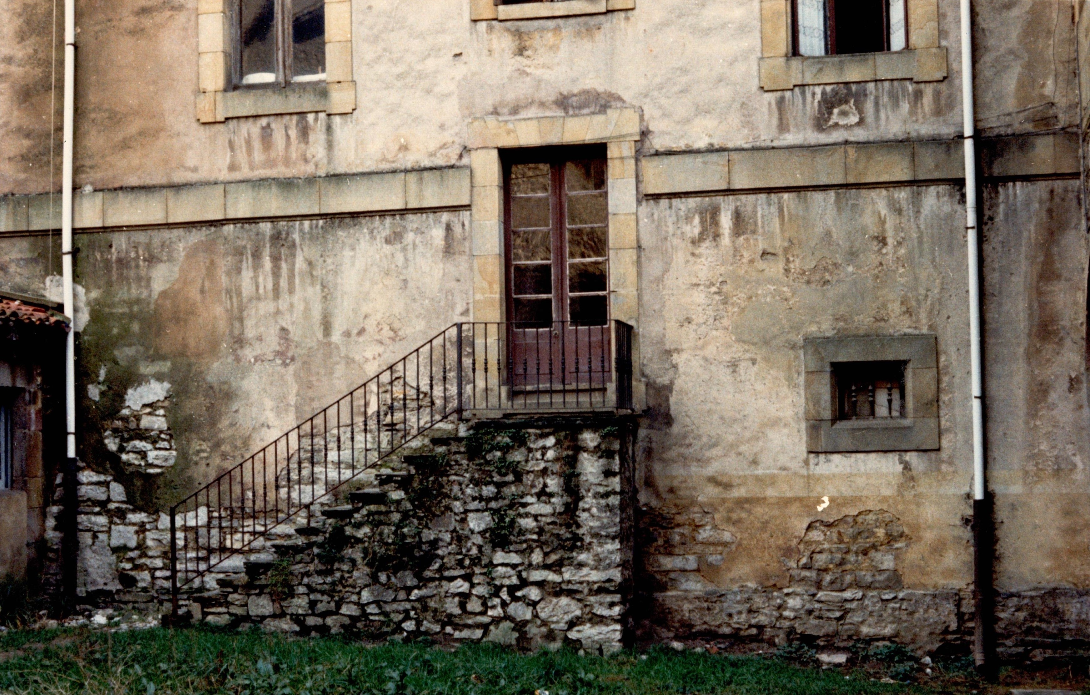 Se ve una parcela con hierba verde oscuro. Hay unas escaleras que comunican este exterior con el interior del edificio, para acceder hay que atravesar una puerta. Se ven dos ventanas de doble hoja y abajo, a la altura de las escaleras una pequeña ventana con tres barrotes. La pared de la fachada interior del edificio está algo deteriorada, tiene manchas, está algo sucia...
