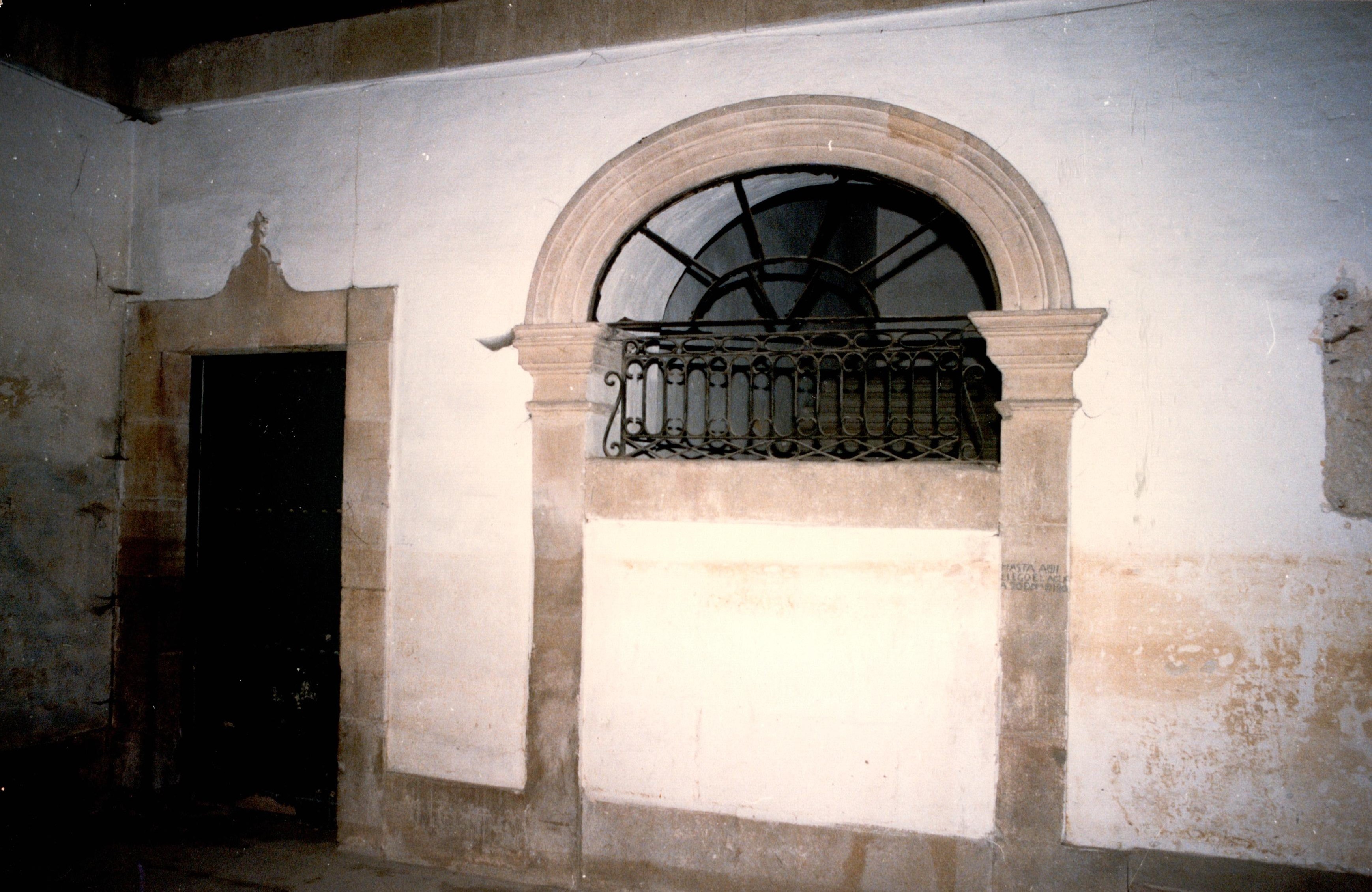 Se ven las paredes blancas y desgastada, algunos elementos decorativos del interior, como arcos y elementos arquitectónicos. A la izquierda hay una puerta. La pared empieza a perder color y hay restos de humedad.