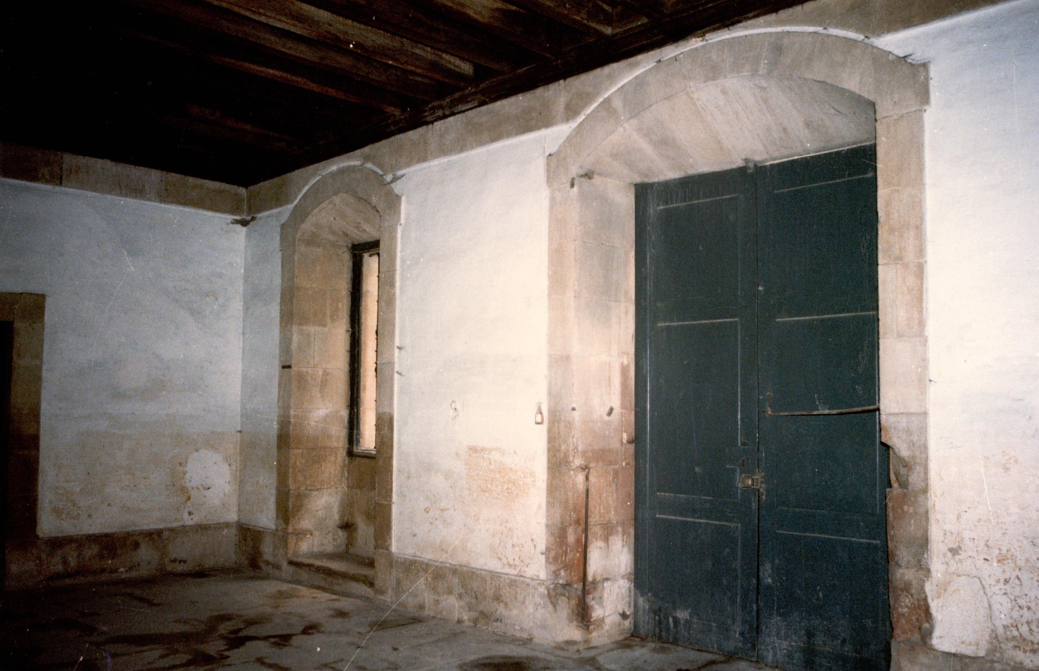 Se ve una puerta de color verde oscuro y a la izquierda una ventana. Es el interior, la planta baja de un edificio. Las paredes son blancas y el techo es de madera.