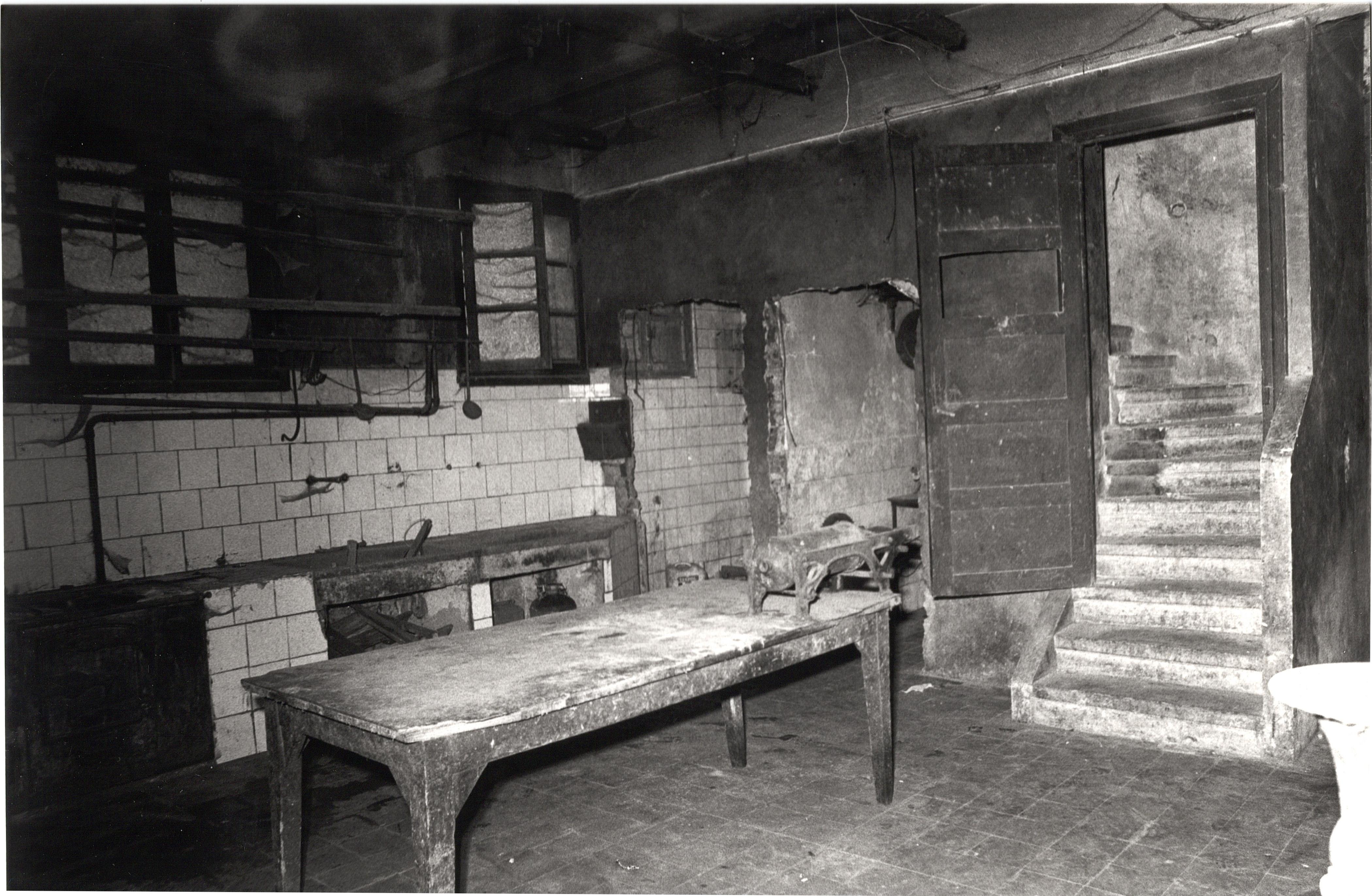 Se ve un espacio, la pared con algunas baldosas, unas encimeras y una mesa con una herramienta encima de la mesa. Al fondo hay como dos puertas que dan acceso a otras estancias y a la derecha de la fotografía hay una puerta y detrás unas escaleras de piedra. 