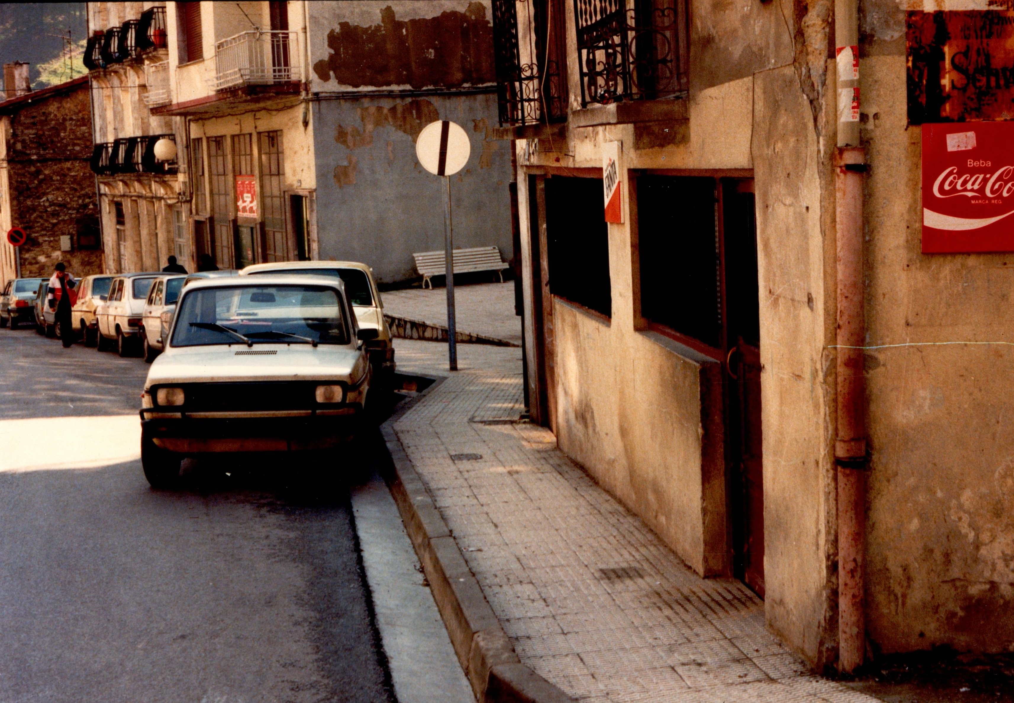 Se ve una calle donde hay aparcados varios coches en fila, en paralelo. Los coches son negros, grises y negros. A la derecha se ven edificios y también en el primero de ellos hay un bar.