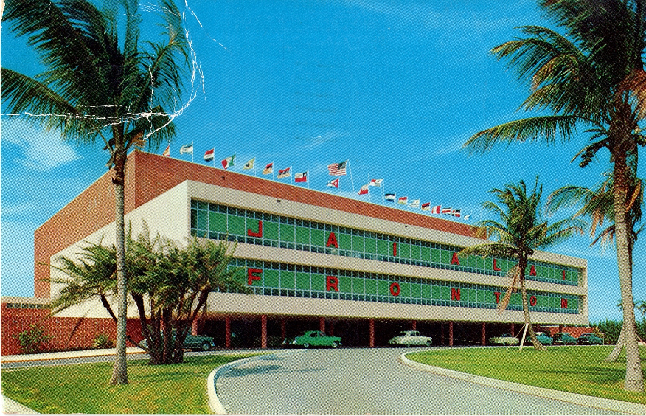 Fotografía de la postal del Jai Alai fronton en la que se ve la parte exterior de acceso a automoviles, y la fachada principal al completo. En el exterior hay jardines y palmeras.