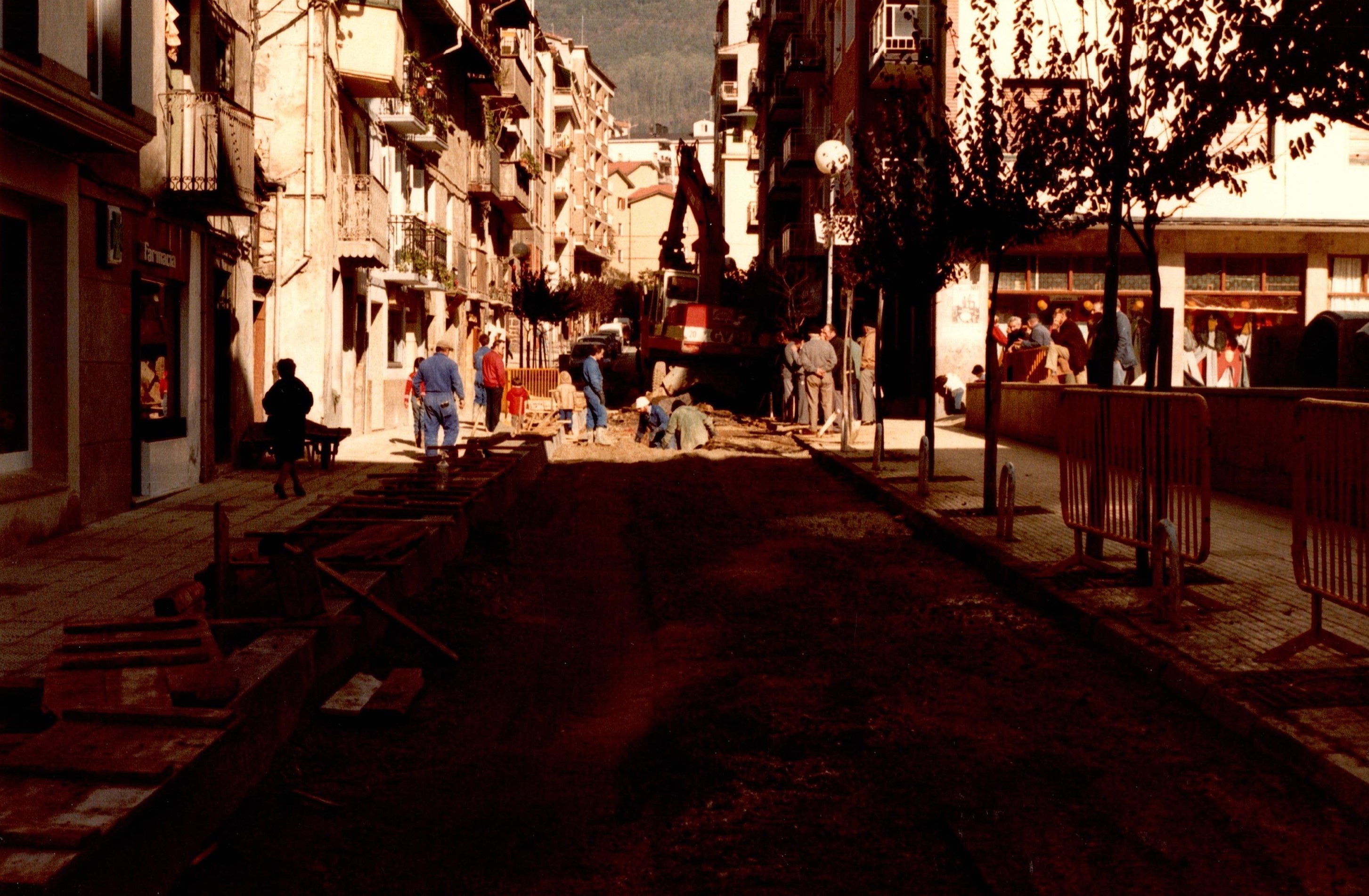 Se muestra la calle Zubiaurre, el primer tramo (el más cercano a quien tomase la fotografía) está muy oscuro porque el sol no entra y está muy sombrío. Aún así se ve el asfalto levantado y que hay obras en ese momento. En la parte que da el sol se ve a algunos obreros trabajando y la maquinaria con la que hacen la obra. A los lados sobre la acera hay gente transitando.