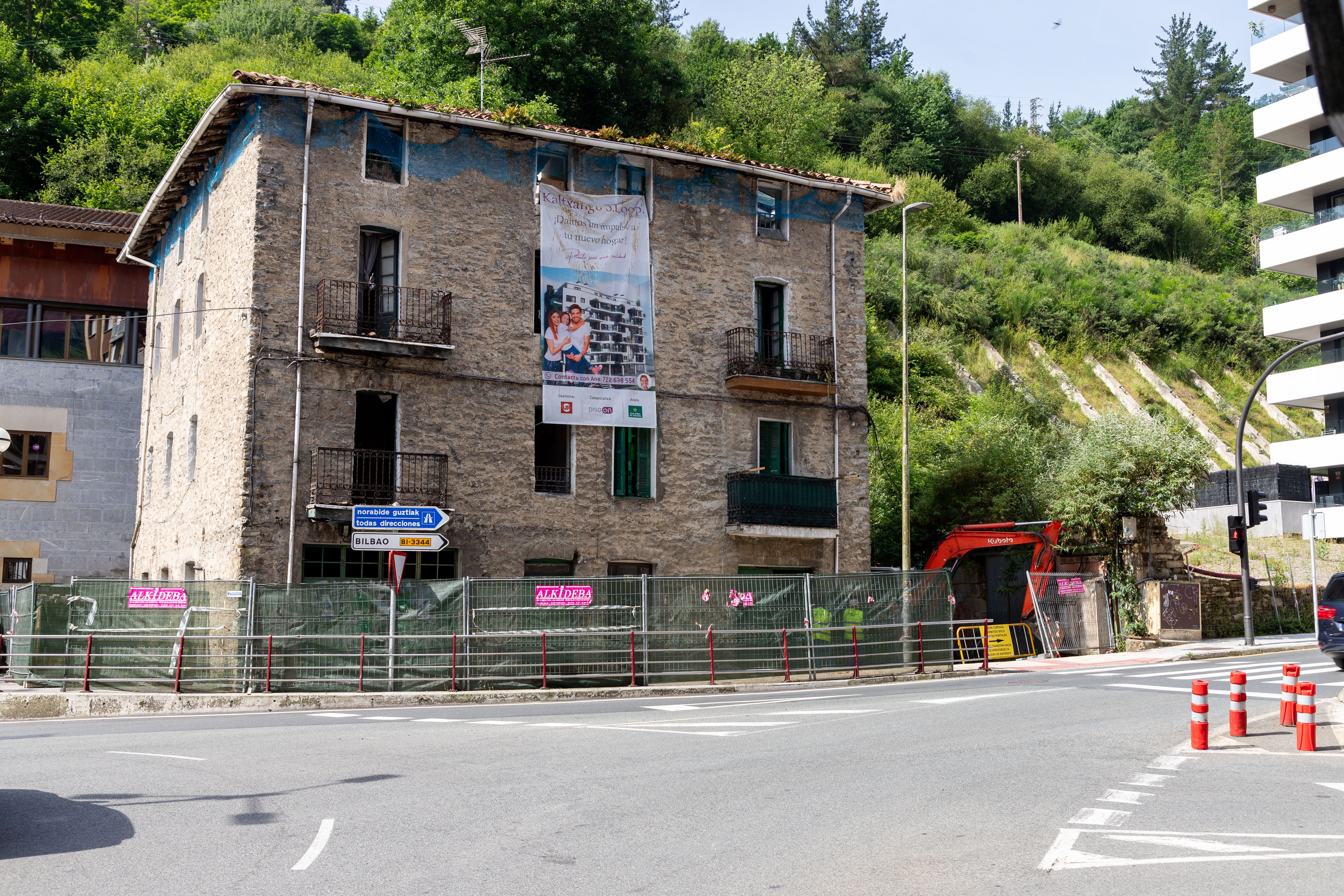 Fachada principal del edificio que van a derribar. El perímetro está delimitado para que los peatones y aquellas personas ajenas a las obras no puedan acceder. Un cartel que cuelga desde la parte superior anuncia la construcción de las viviendas que se van a construir tras el derribo. El edificio tiene cuatro plantas y es grande, de piedra y con cuatro balcones individuales en la fachada principal. Hay dos puertas de acceso. Algunas ventanas están rotas. A la derecha se ve parte de la maquinaria.
