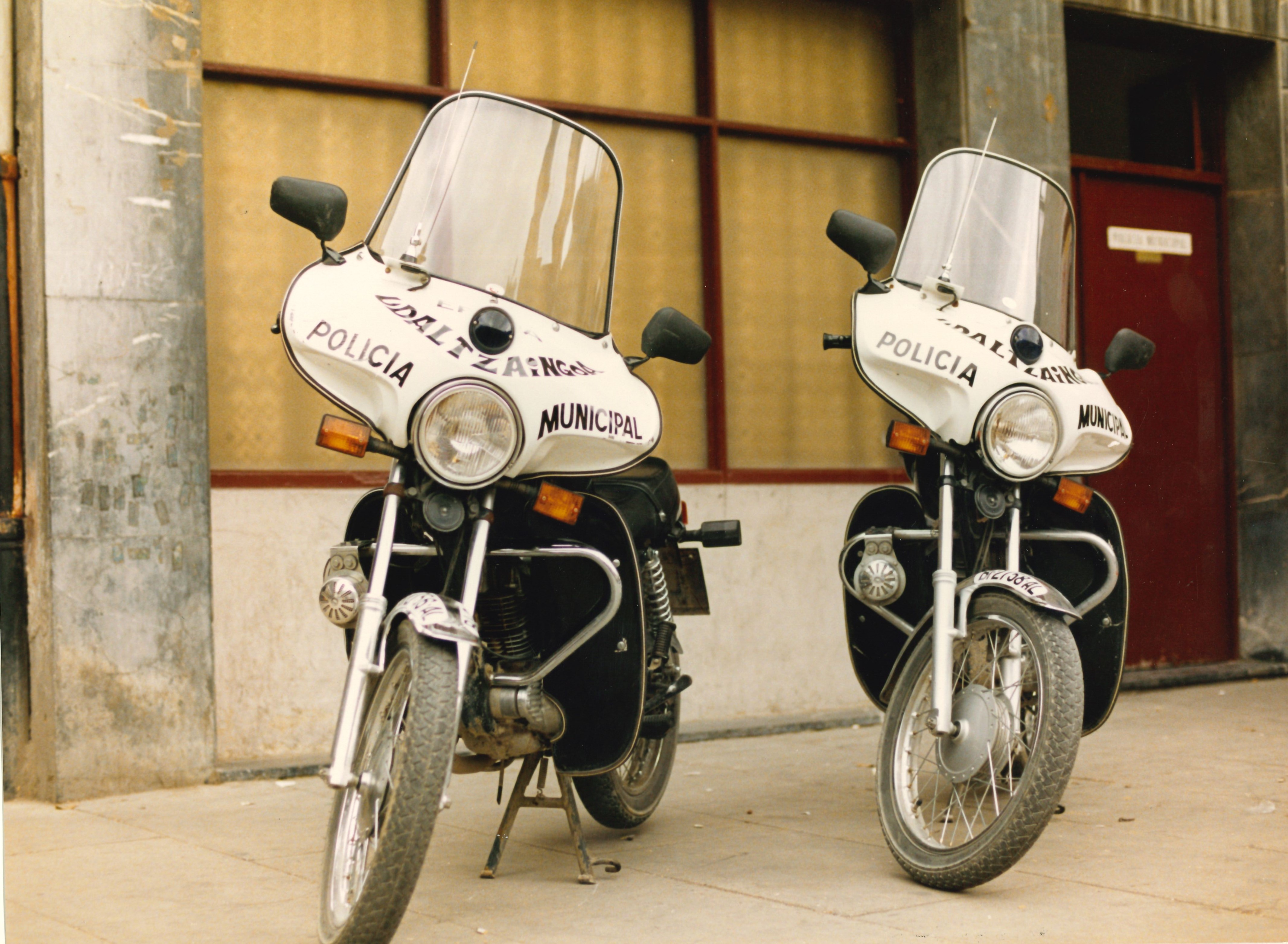 Dos motocicletas de la policía municipal de Ermua, son blancas y negras. La parte delantera alrededor del foco central y debajo de la pantalla de la moto es blanca y en letras negras se puede leer "policía municipal - udaltzaingoa. Las ruedas son altas y finas, tienen guardarbarros, dos retrovisores y dos indicadores delanteros de luz naranja. Están apoyadas sobre la pata de cabra que tiene cada motocicleta.