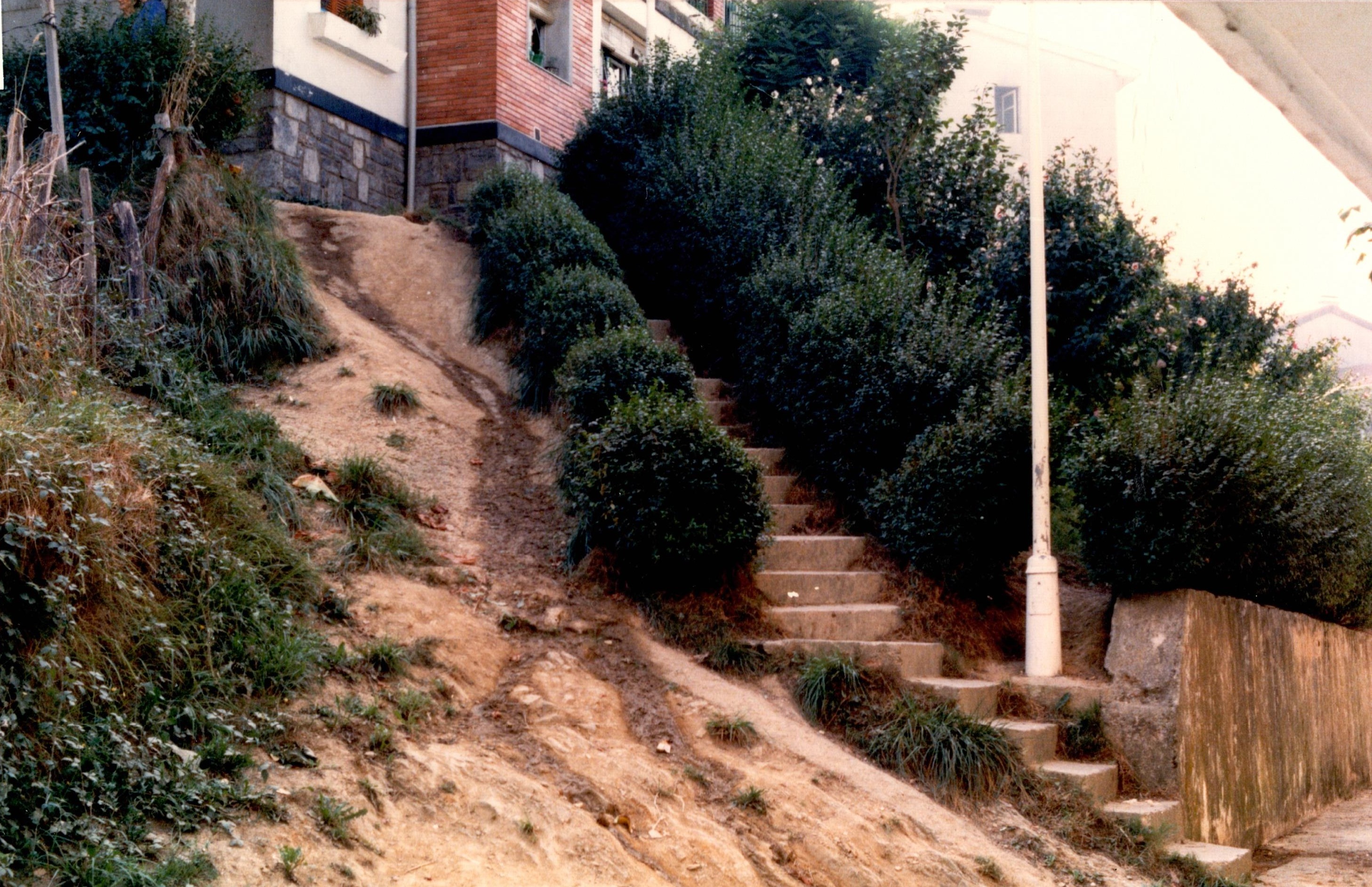 Se muestra un acceso en ascensión a unas viviendas en el barrio de Santa Ana. Solo hay un tramo estrecho de escaleras sin barandilla y a su izquierda una rampa sin trabajar de barro. Hay maleza y arbustos. También se ve una farola blanca. 