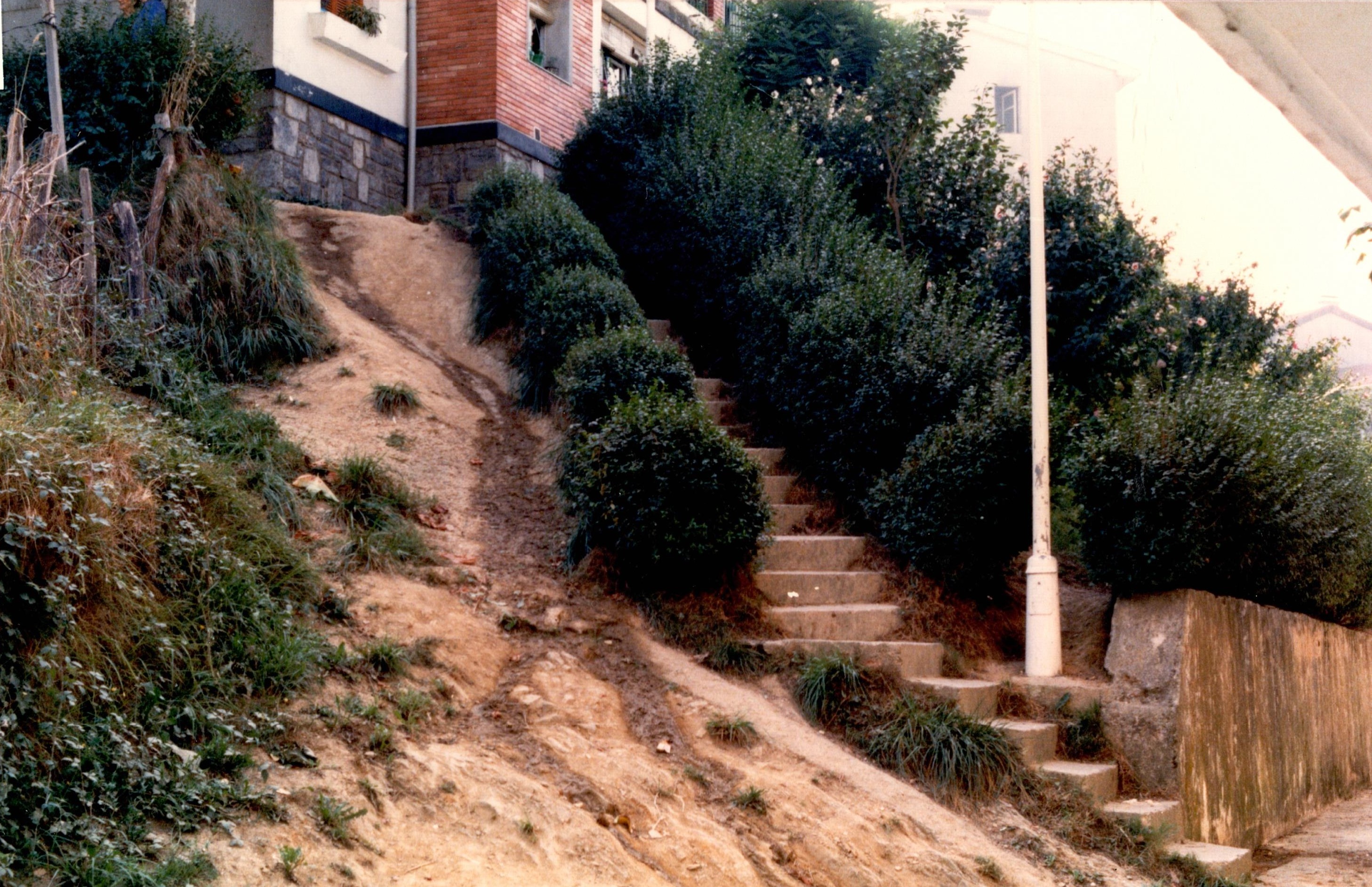 Se muestra un acceso en ascensión a unas viviendas en el barrio de Santa Ana. Solo hay un tramo estrecho de escaleras sin barandilla y a su izquierda una rampa sin trabajar de barro. Hay maleza y arbustos. También se ve una farola blanca.