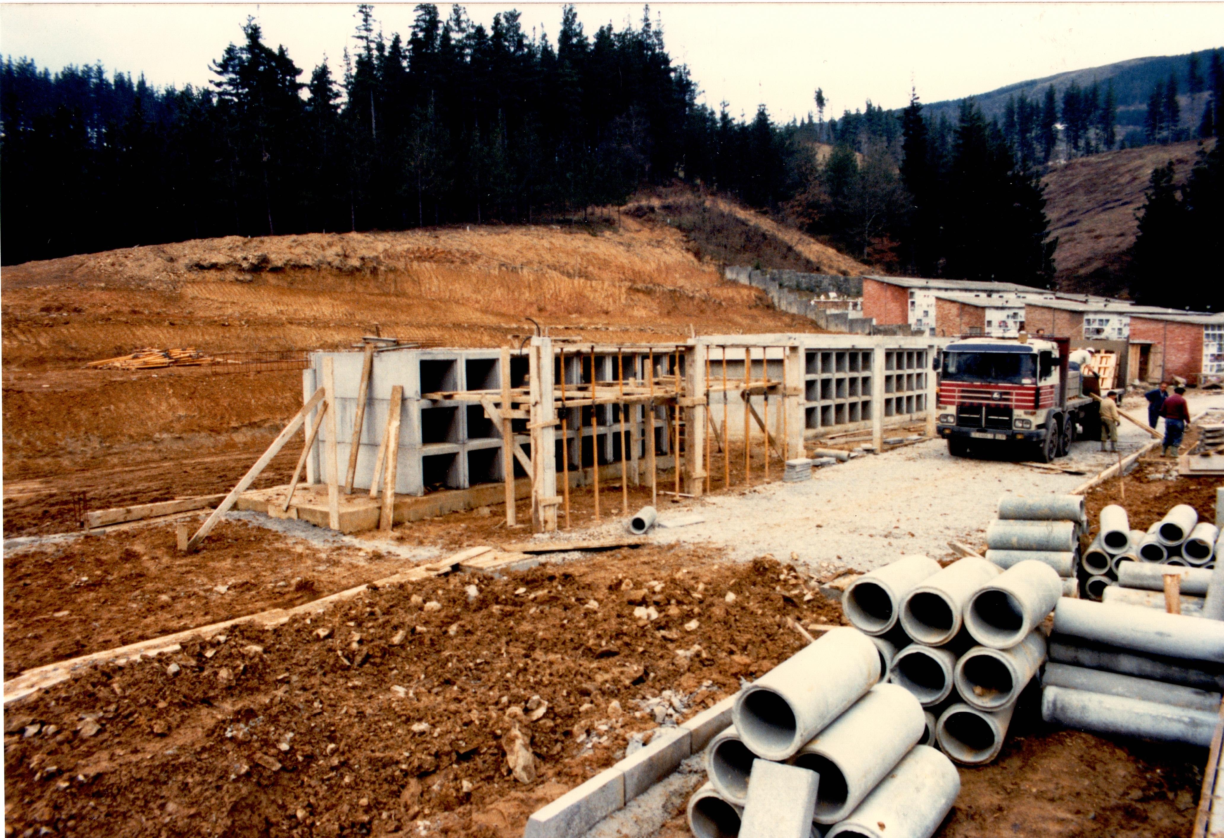 Se ven los nichos de lo que será el cementerio construyéndose, hay material de obra como unos tubos de cemento, vigas de madera, barras de metal... también se ve un camión de obra del que unos obreros están bajando material. Encima del camión hay un obrero, otro le ayuda y hay dos fuera del camión. La tierra de alrededor está levantada, es marrón oscura en algunas zonas y marrón más clara en otras.
