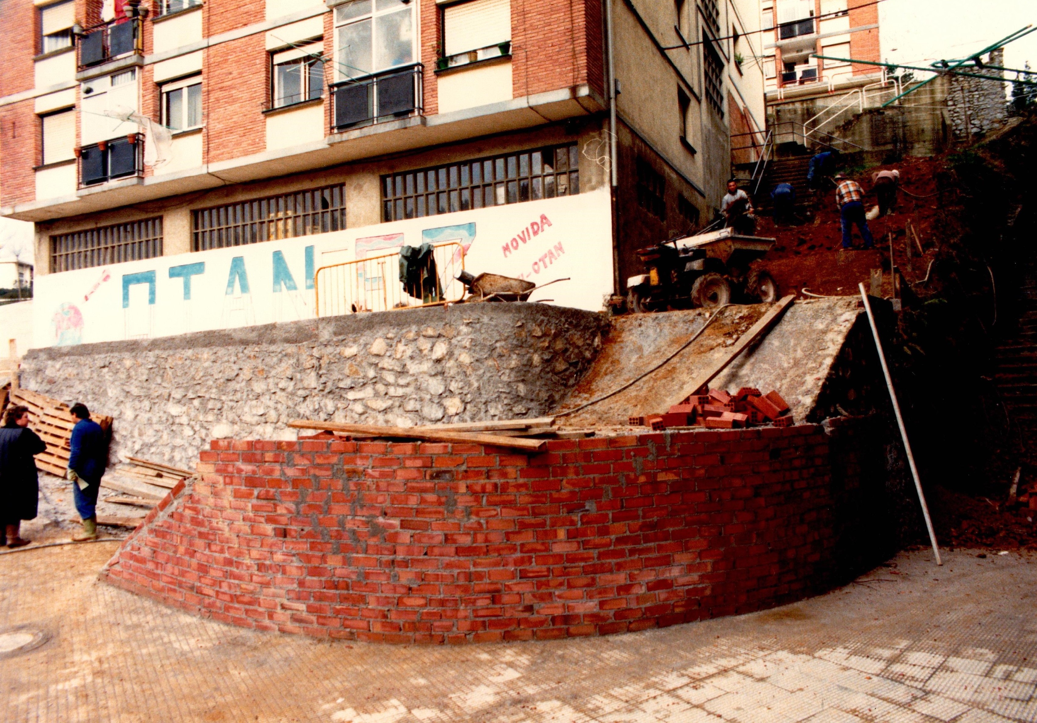 Se ve parte de una estructura sin acabar hecha con ladrillos rojos y cemento gris, todavía fresco parece. Detrás hay un edificio de viviendas, en la parte inferior de ese edificio hay una pintada que dice: OTAN - EZ movida. A la izquierda hay dos personas hablando, una de ellas va con buzo azul y botas verdes de trabajo. A la derecha, en las obras de accesibilidad hay varios obreros trabajando y maquinaria de trabajo.