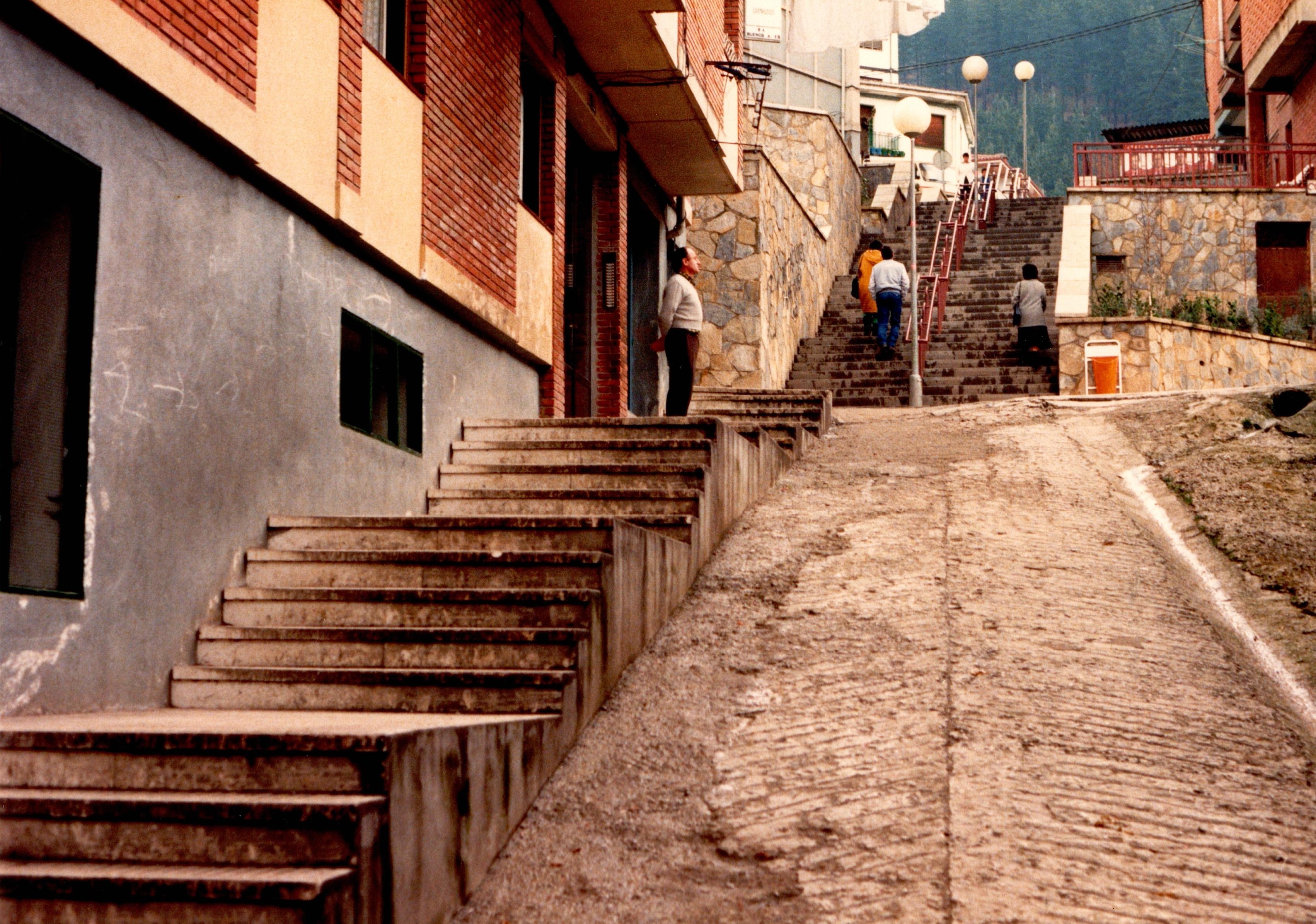 Se muestra la subida de una cuesta y unas escaleras. A la izquierda, a pie de calle hay viviendas pegadas a las escaleras. El asfalto está en malas condiciones, sobre todo la cuesta. Hay un señor en uno de los tramos de las escaleras de pie, quieto y otras tres personas subiendo por otro tamo de escaleras. 